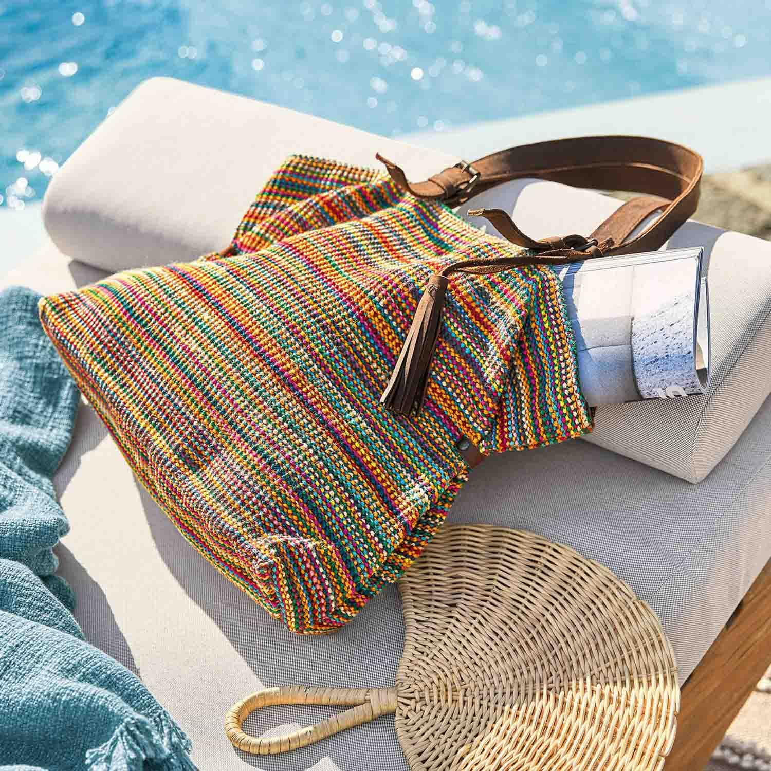 Sac tissé multicolore avec bandoulière en cuir marron posé sur un coussin clair près de la piscine