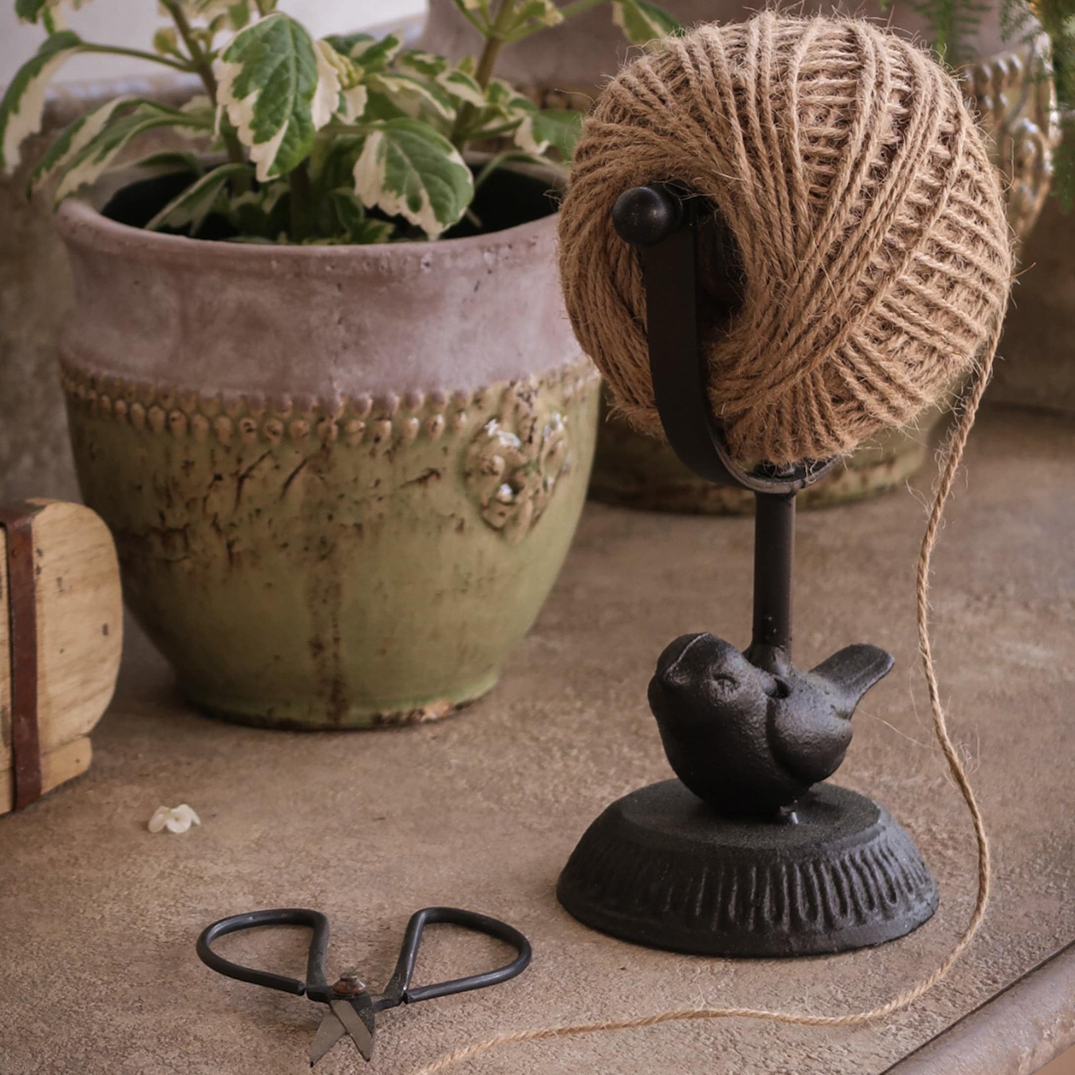 Support en métal noir avec pelote de ficelle et décoration oiseau devant un pot de fleurs vert
