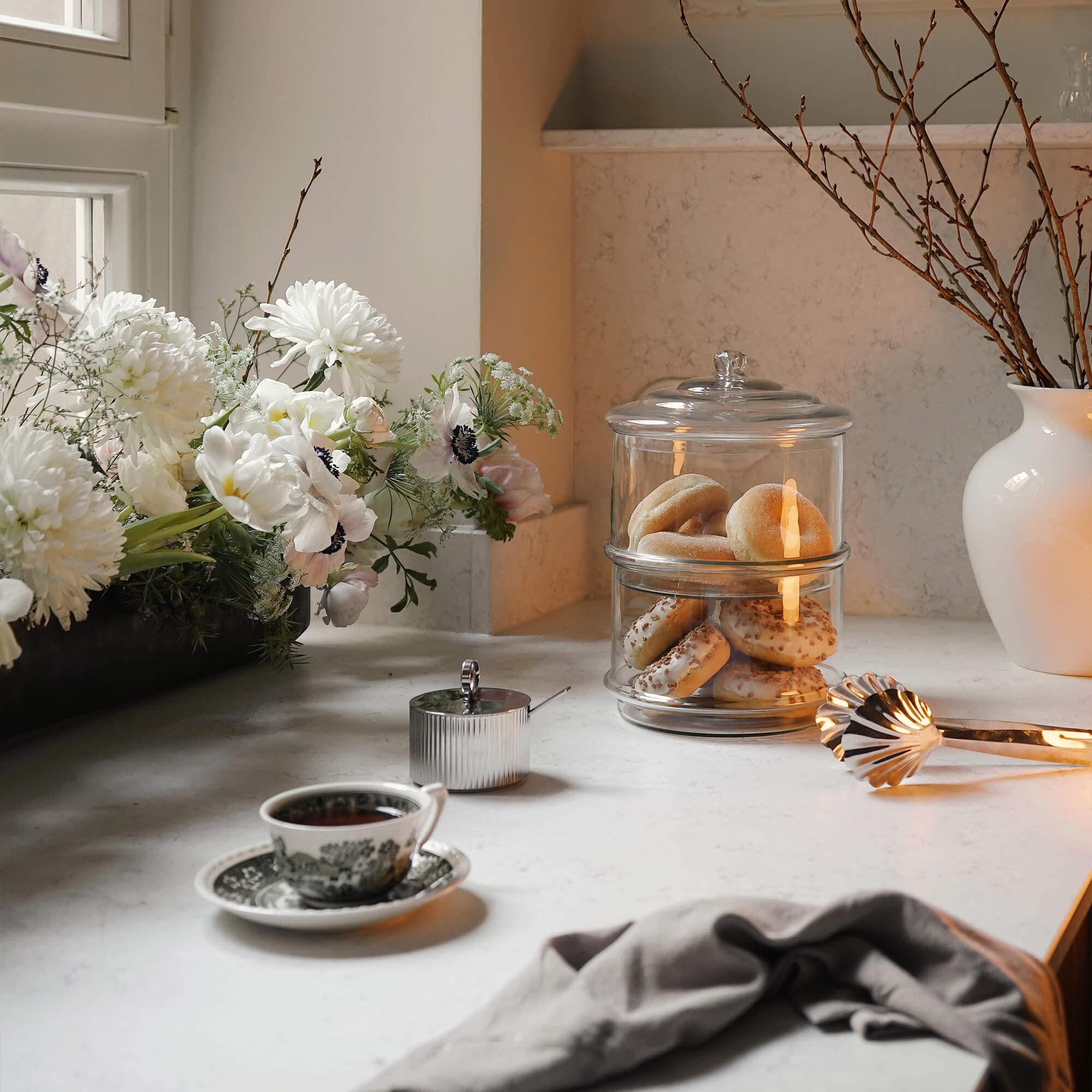 Boîte à biscuits en verre à deux étages sur un plan de travail clair avec fleurs blanches et tasse de thé