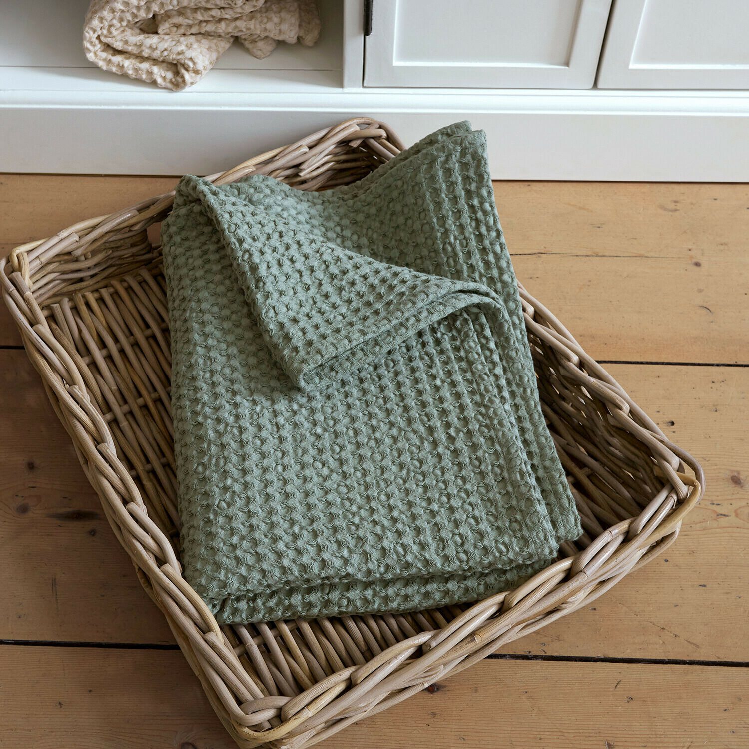 Serviette de bain Maralei Plaid vert en coton gaufré dans un panier en osier posé sur un sol en bois clair