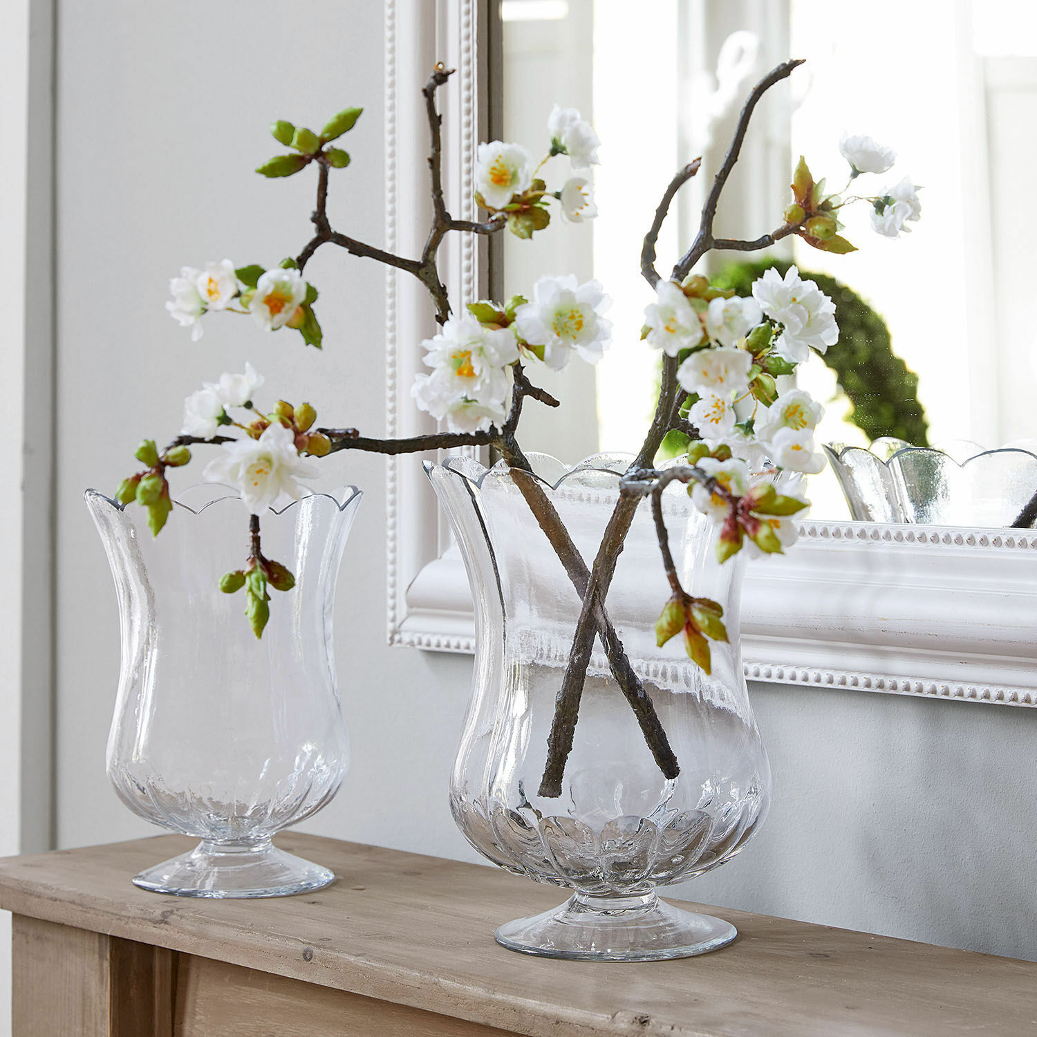 Lot de 2 fleurs décoratives Gráda Vases en verre transparent avec bord ondulé sur table en bois décorés de fleurs blanches artificielles