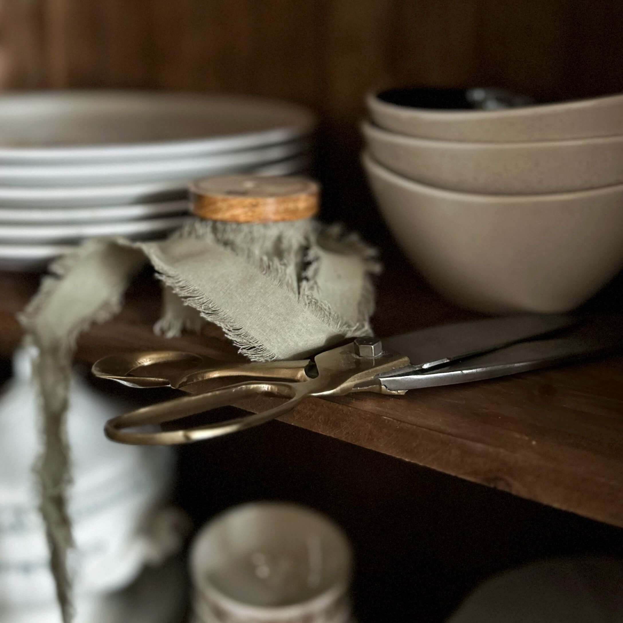Ciseaux en laiton anciens sur une étagère en bois près de bols et assiettes beige dans une cuisine
