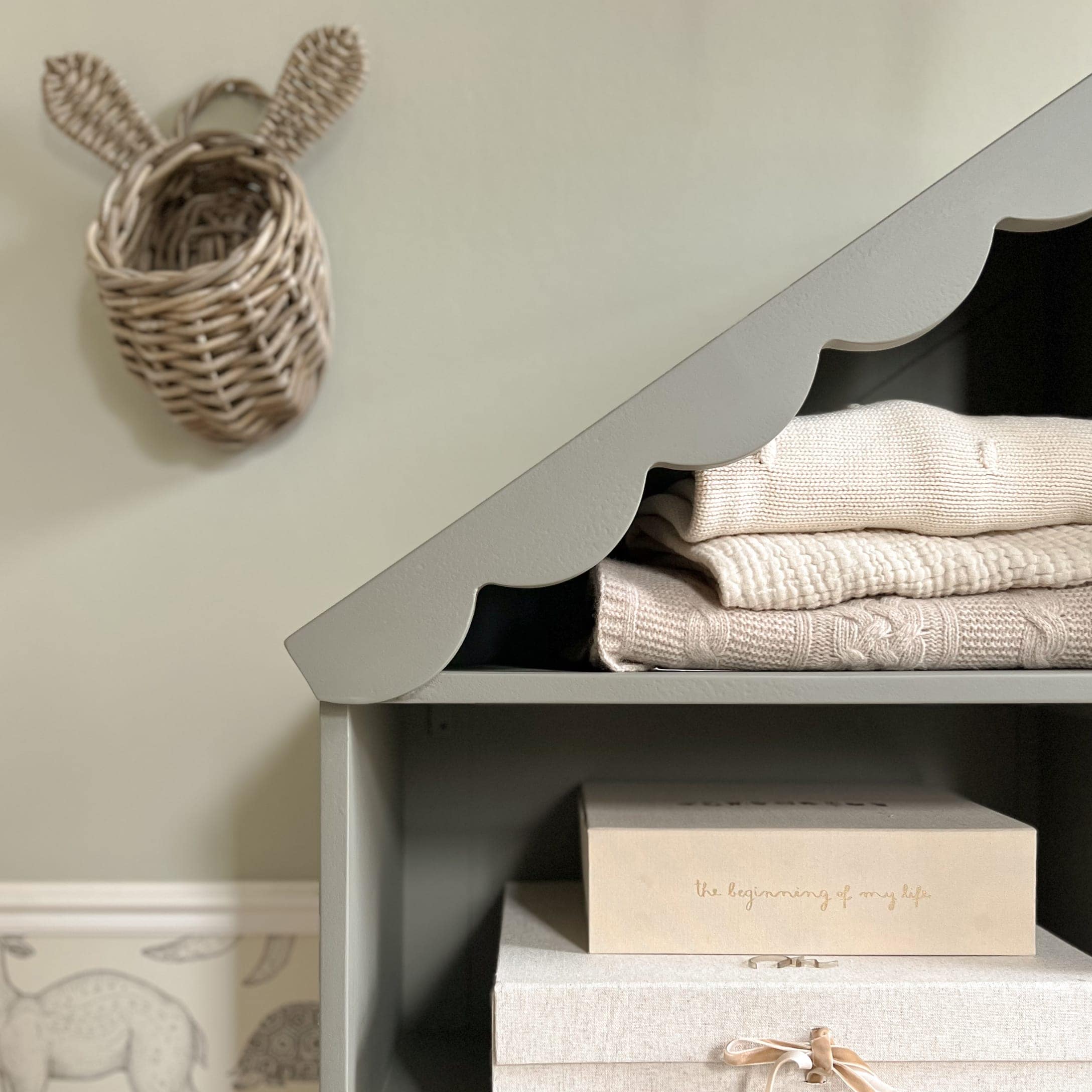 Étagère grise en bois avec bord ondulé, pulls en tricot crème et boîtes de rangement beige clair