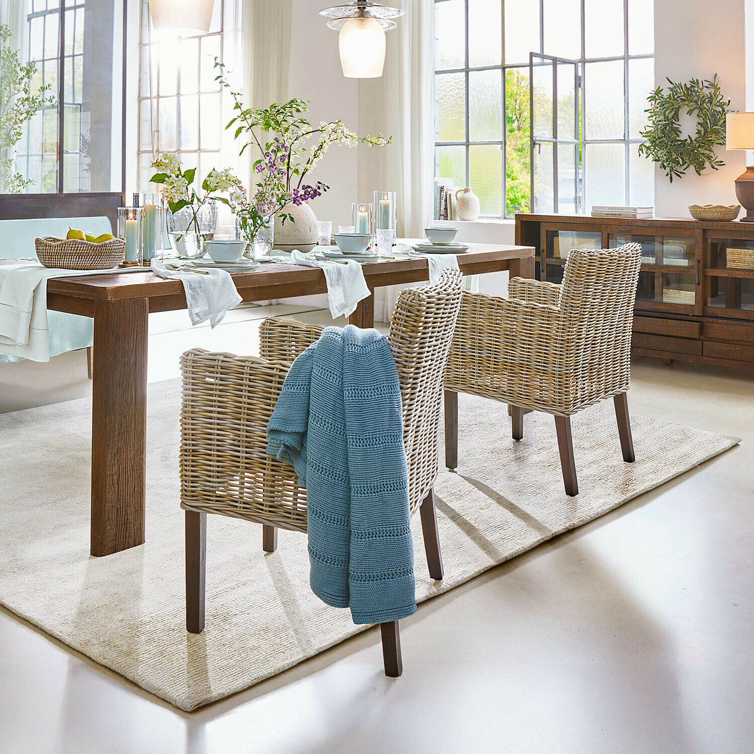 Fauteuil Forêt Douce Salle à manger avec table en bois et chaises en rotin naturel, décorations florales et plaid bleu