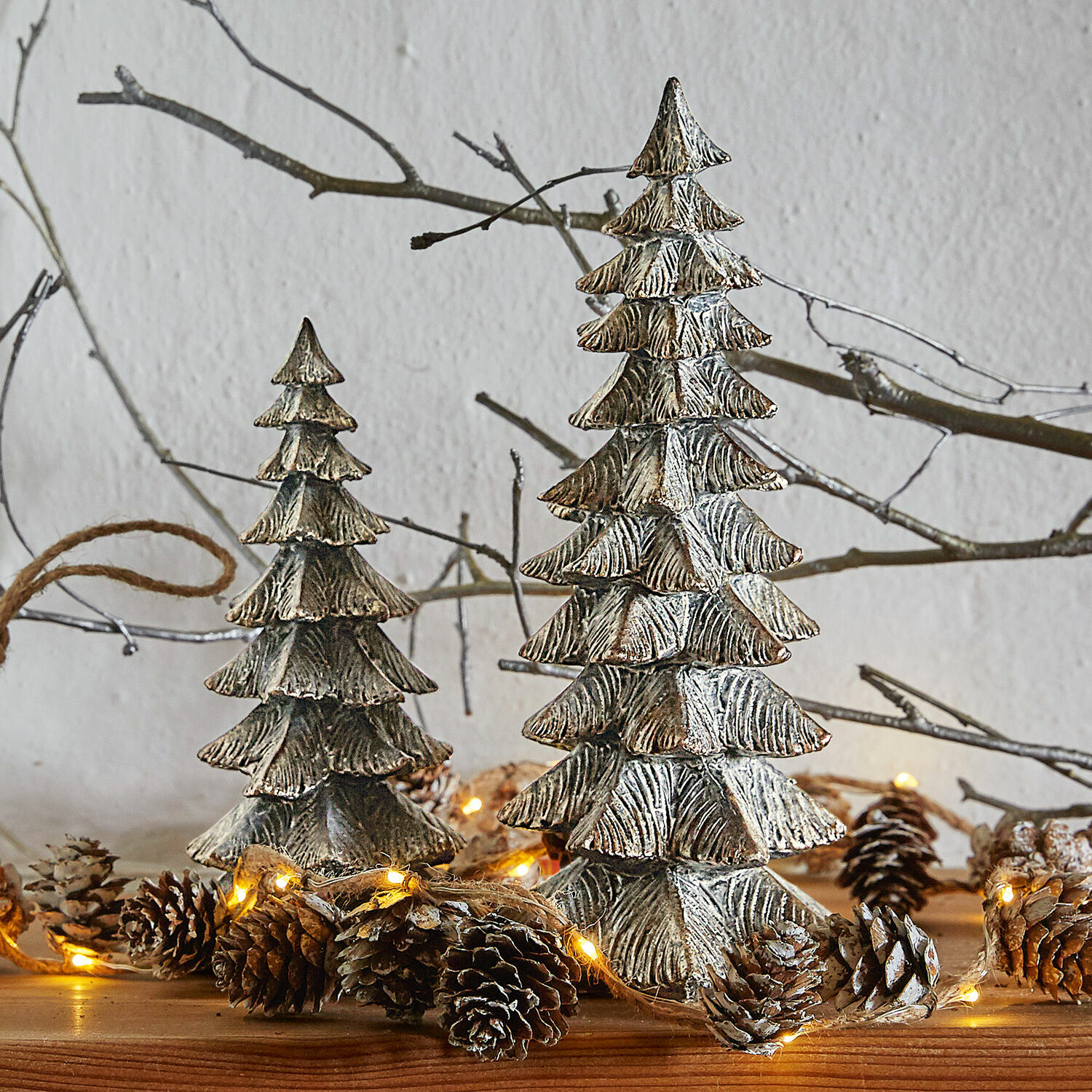 Lot de 2 sapins décoratifs Calavi Décorations sapins en métal argenté sur table en bois avec guirlande lumineuse et pommes de pin