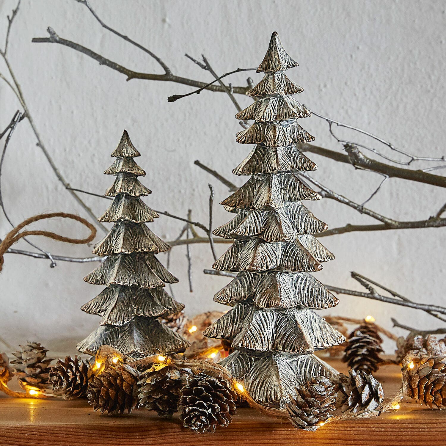 Lot de 2 sapins décoratifs Calavi Décorations sapins en métal argenté sur table en bois avec guirlande lumineuse et pommes de pin