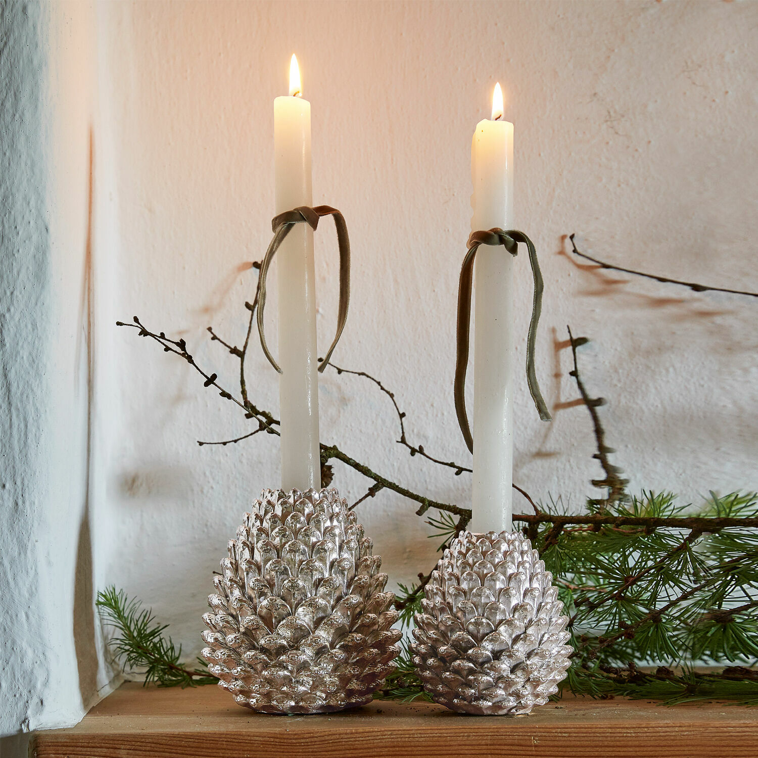 Lot de 2 porte-bougies Mirbells Deux bougeoirs argentés en forme de pomme de pin avec bougies blanches sur une étagère en bois décorée de branches vertes