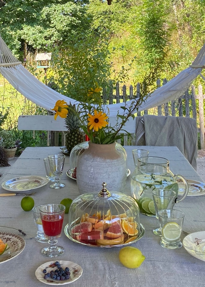 Table de jardin avec nappe grise, fruits et vase à fleurs jaunes devant un hamac blanc et un jardin verdoyant