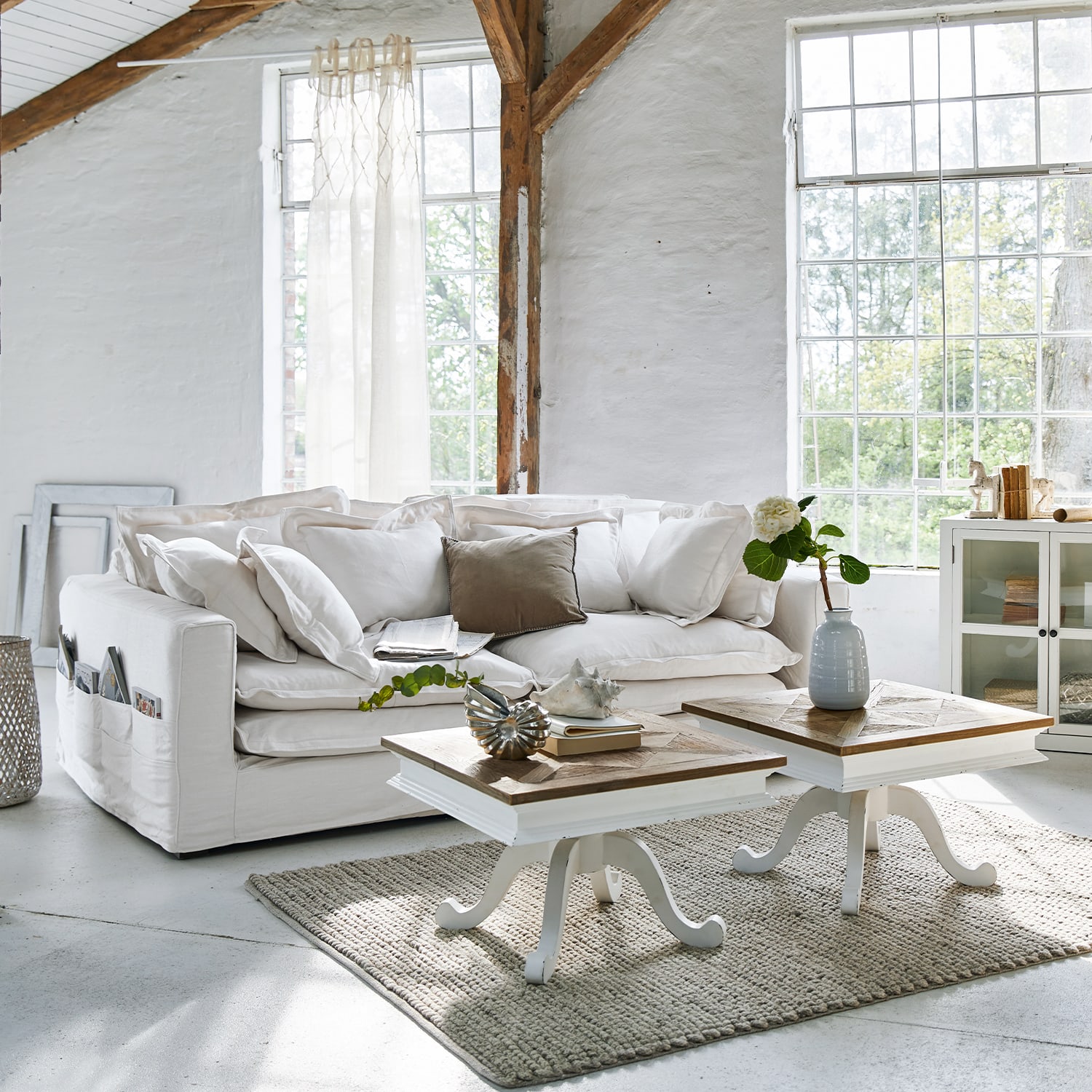 Canapé blanc en coton avec coussins et deux tables basses en bois sur tapis gris dans un salon lumineux