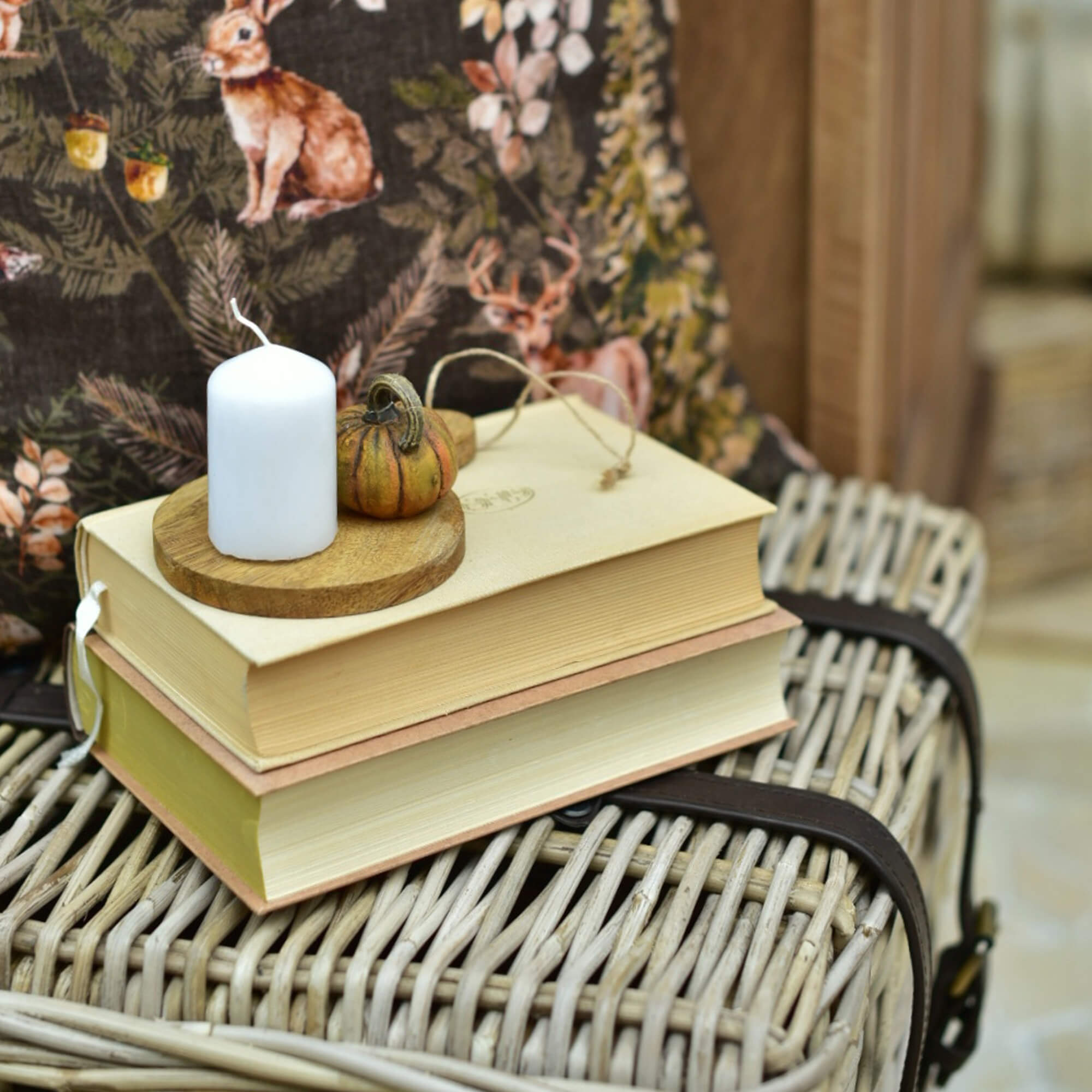 Bougie blanche et petite citrouille décorative sur une planche en bois posée sur des livres empilés sur une valise en osier