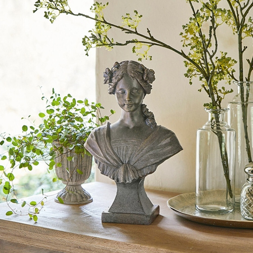 Buste féminin en pierre grise avec fleurs dans les cheveux sur une table en bois décorée de plantes