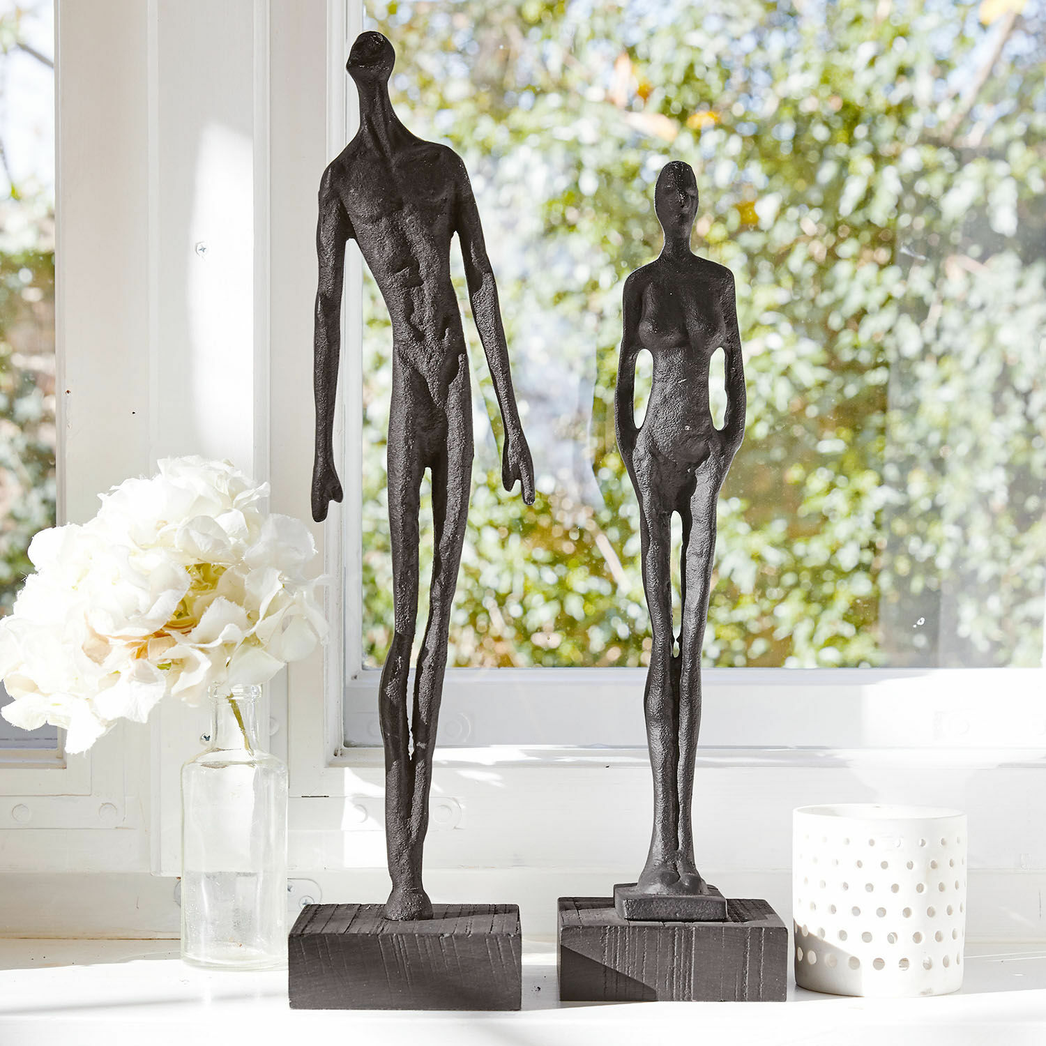 Lot de 2 figurines Perce Deux sculptures noires en métal de figures humaines sur socles en bois devant une fenêtre lumineuse
