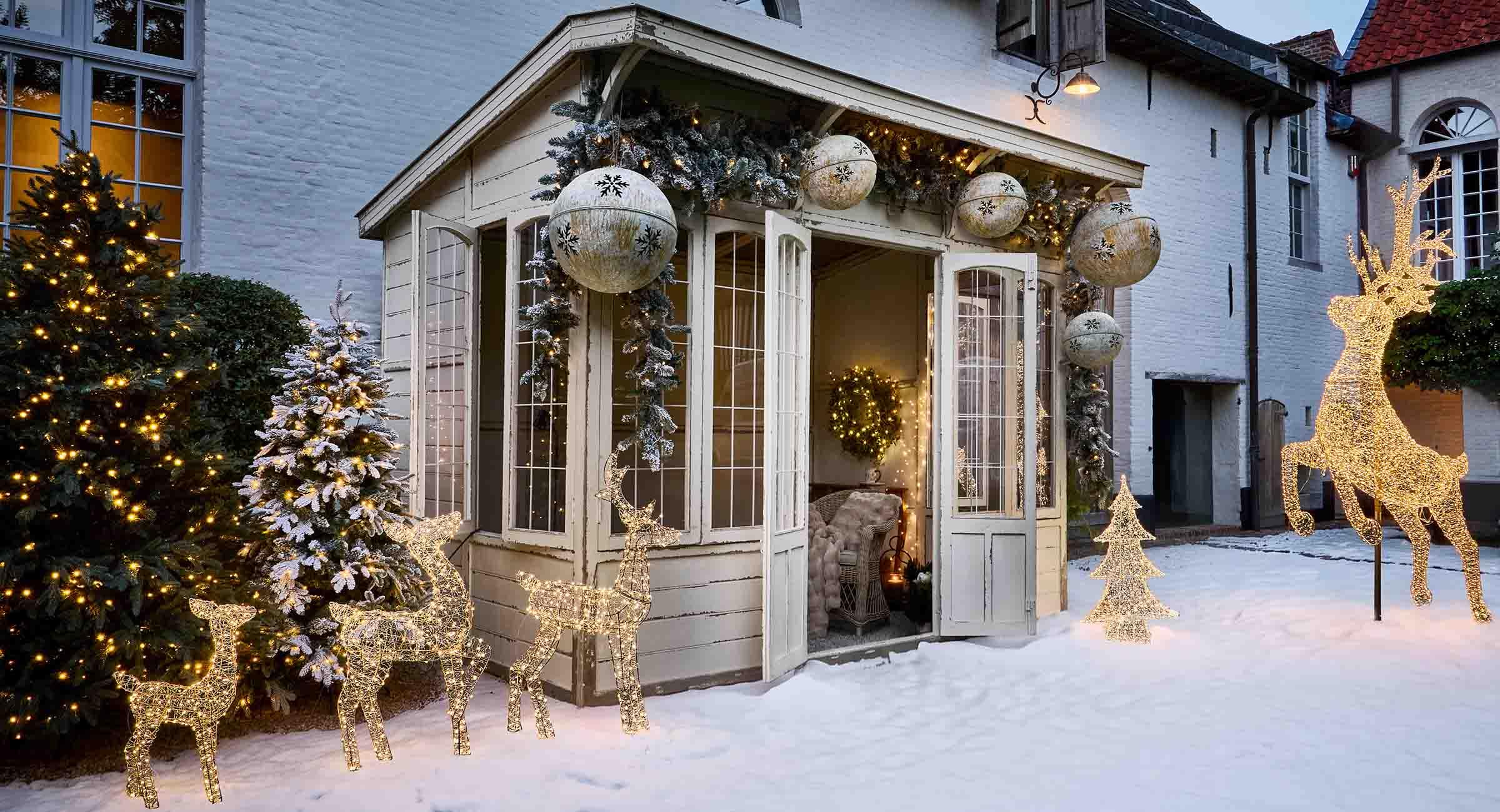 Décorations lumineuses de rennes et sapins sur la neige devant une maison blanche avec ambiance hivernale