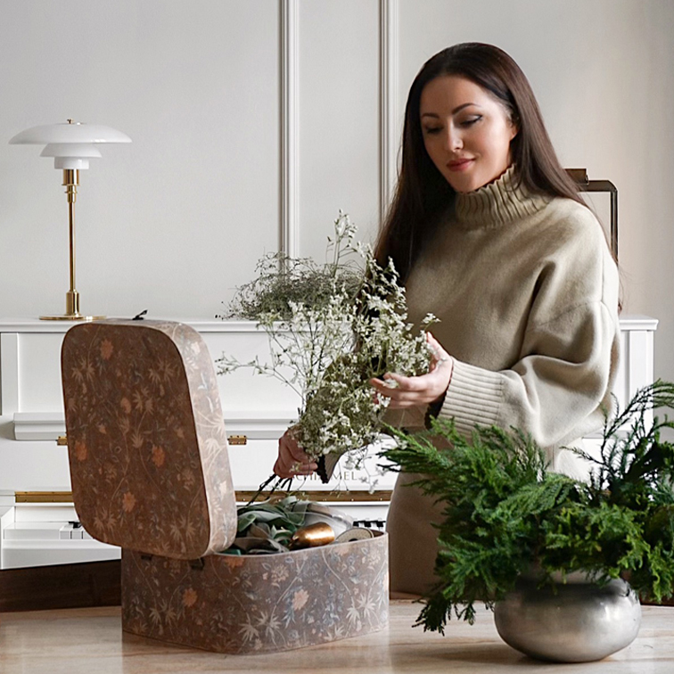 Femme en pull beige tenant un bouquet de fleurs près d'une boîte décorative et d'une plante verte sur la table