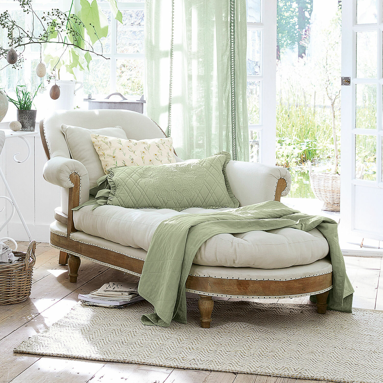 Méridienne Queyranne Chaise longue élégante en bois clair et tissu crème avec couverture verte et coussins dans un intérieur lumineux