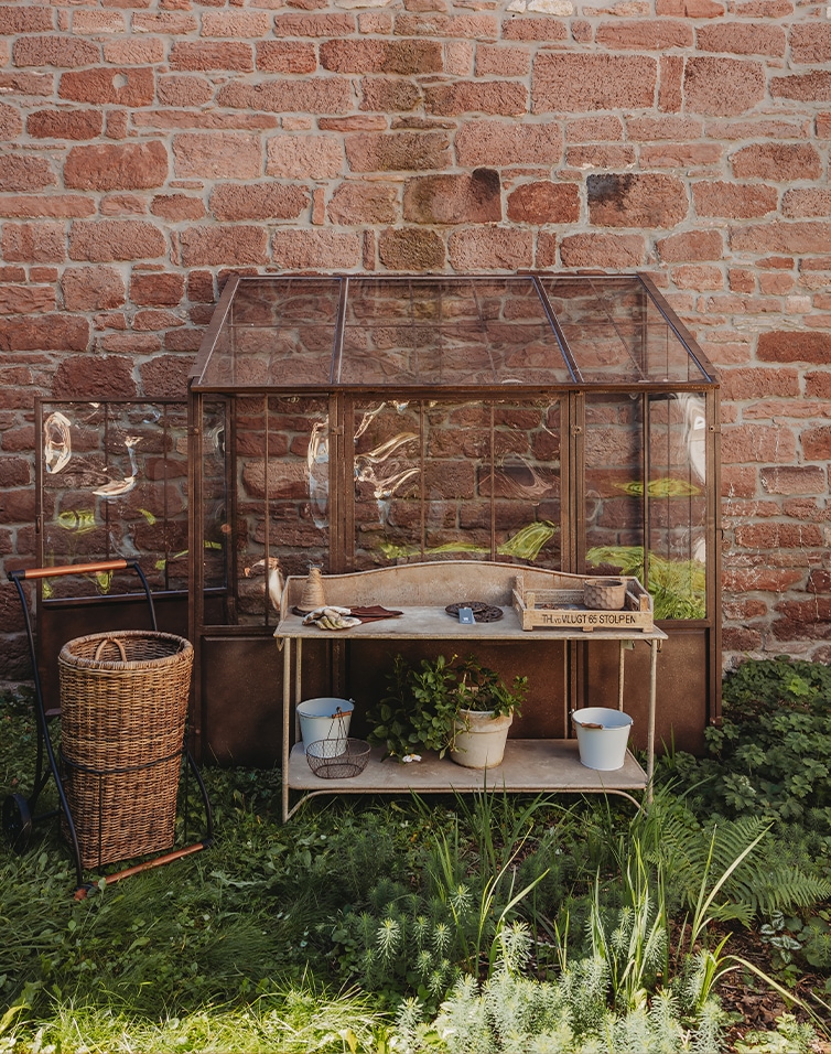 Serre de jardin en verre avec cadre métallique et table en bois devant un mur en briques rouges
