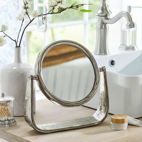 Miroir rond sur support en métal argenté posé sur un plan de salle de bain en bois clair