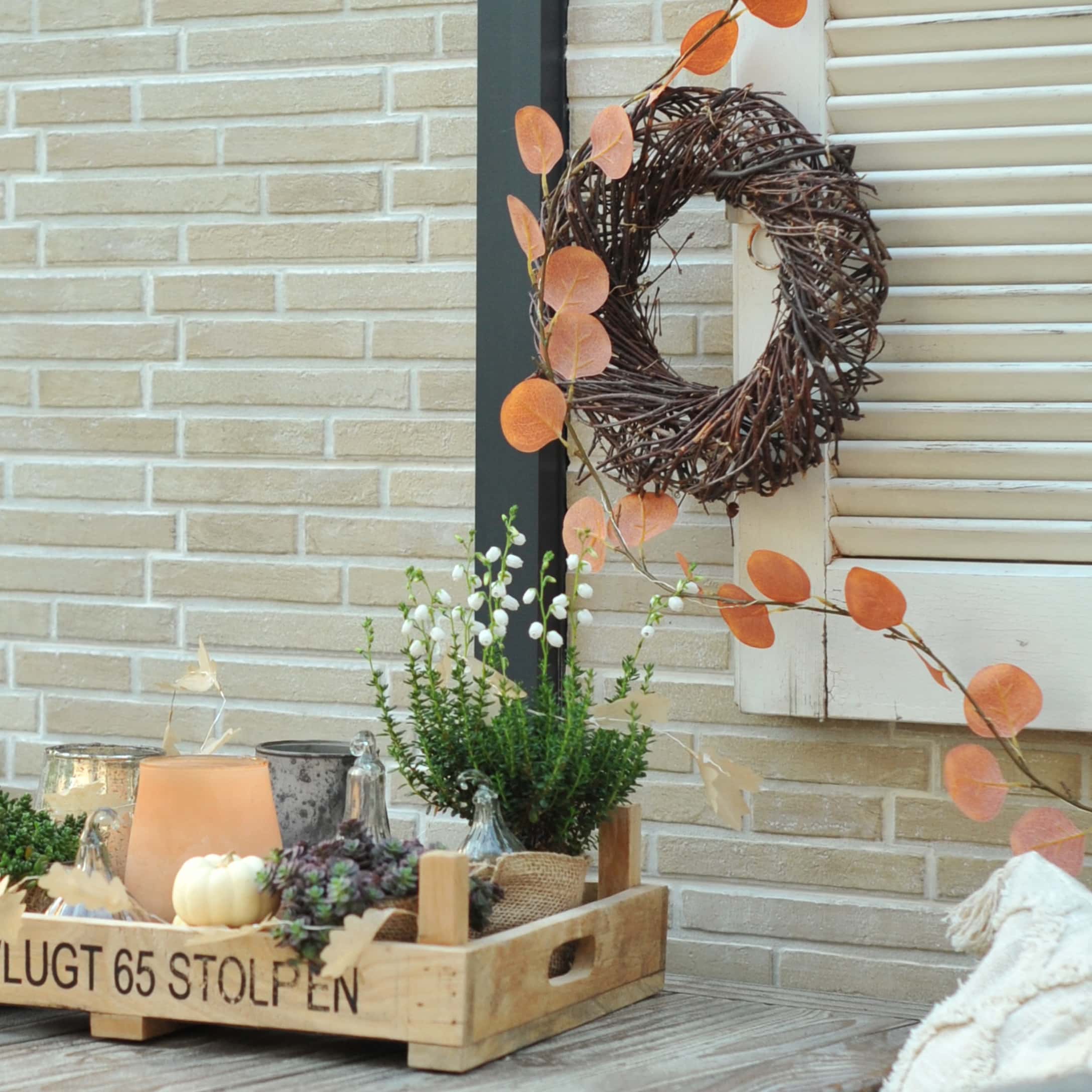 Plateau en bois avec décoration automnale de plantes et bougies devant un mur en briques et volets blancs