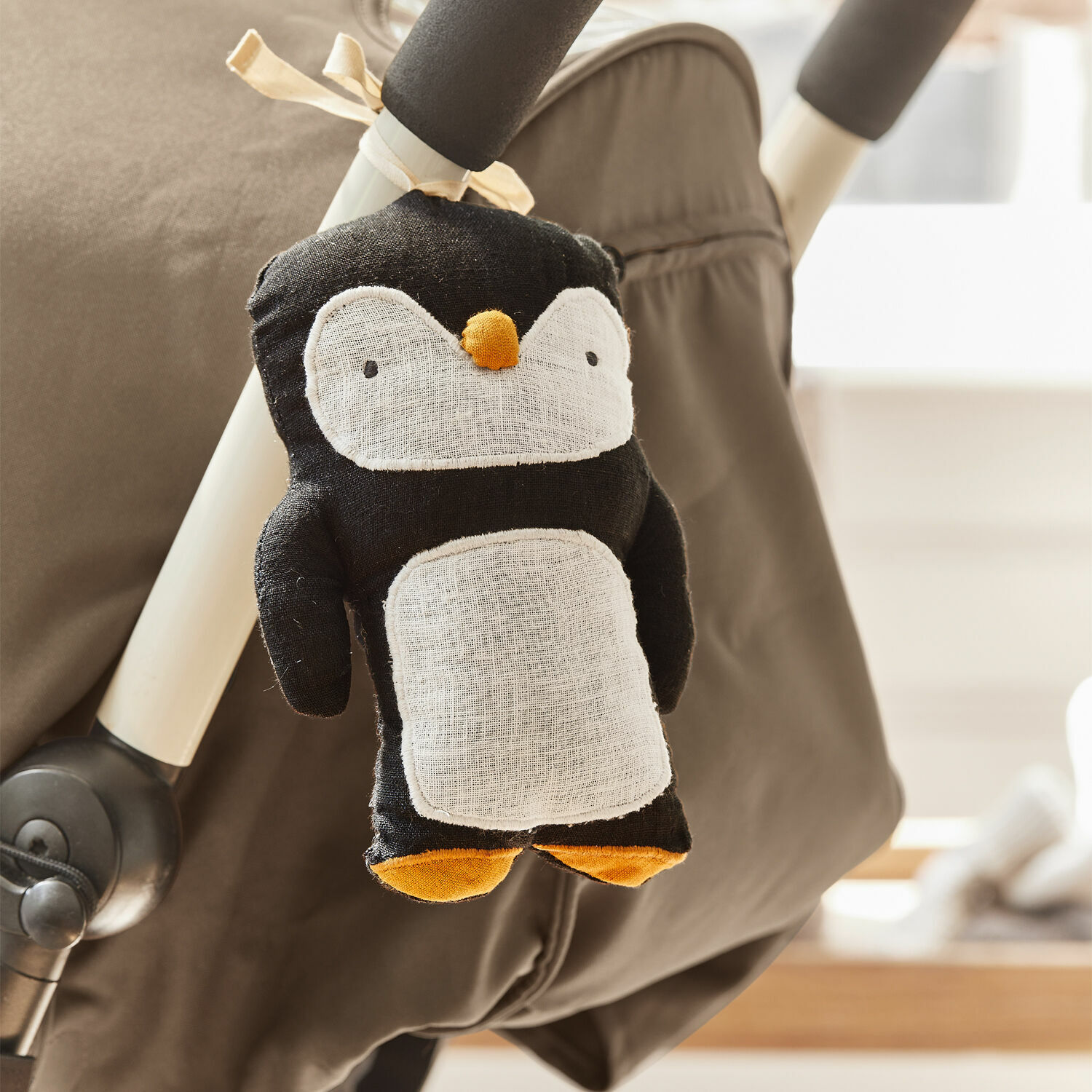 Suspension pour poussette Jocienne Jouet en peluche pingouin noir avec ventre blanc et pieds orange attaché à une poussette