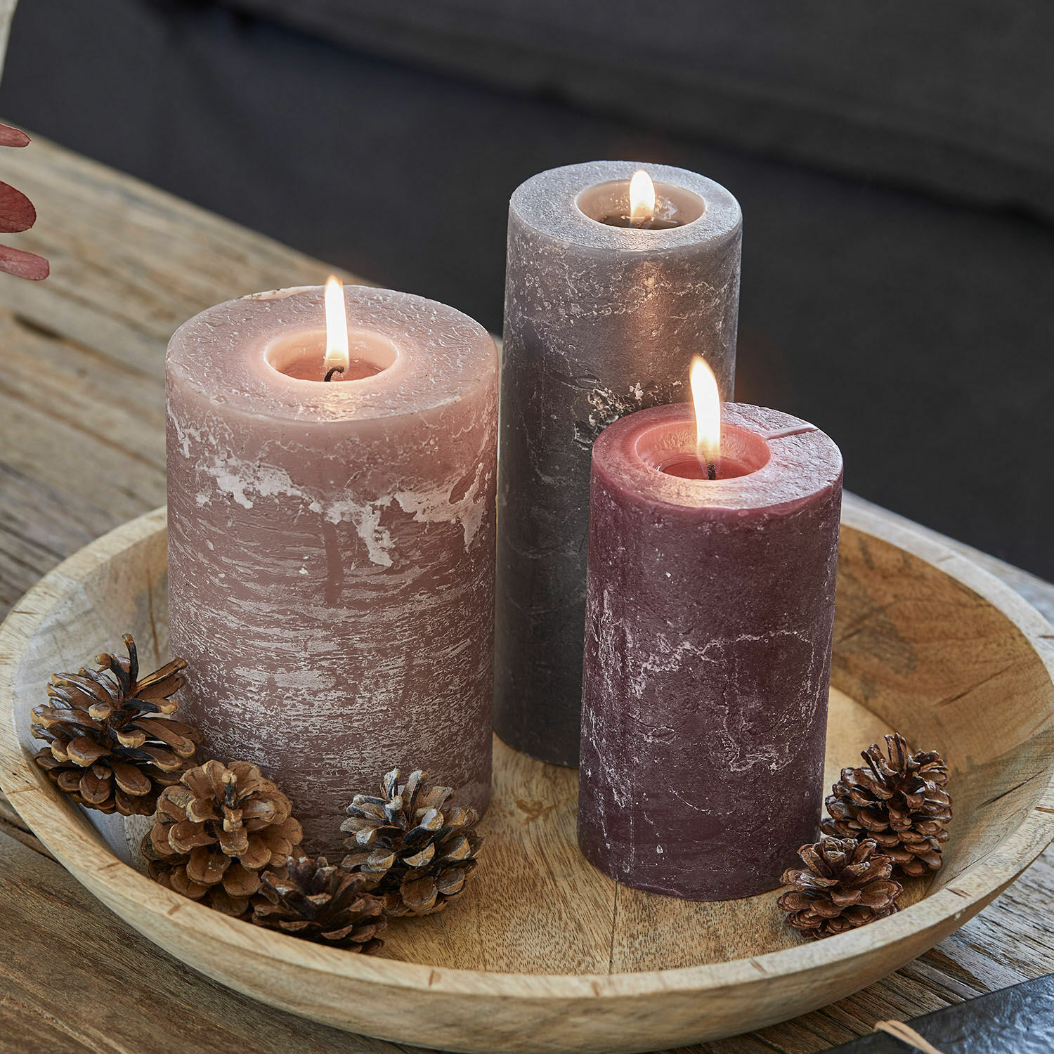 Lot de 3 bougies Pretin Trois bougies allumées en nuances de rose et gris sur un plateau en bois avec des pommes de pin