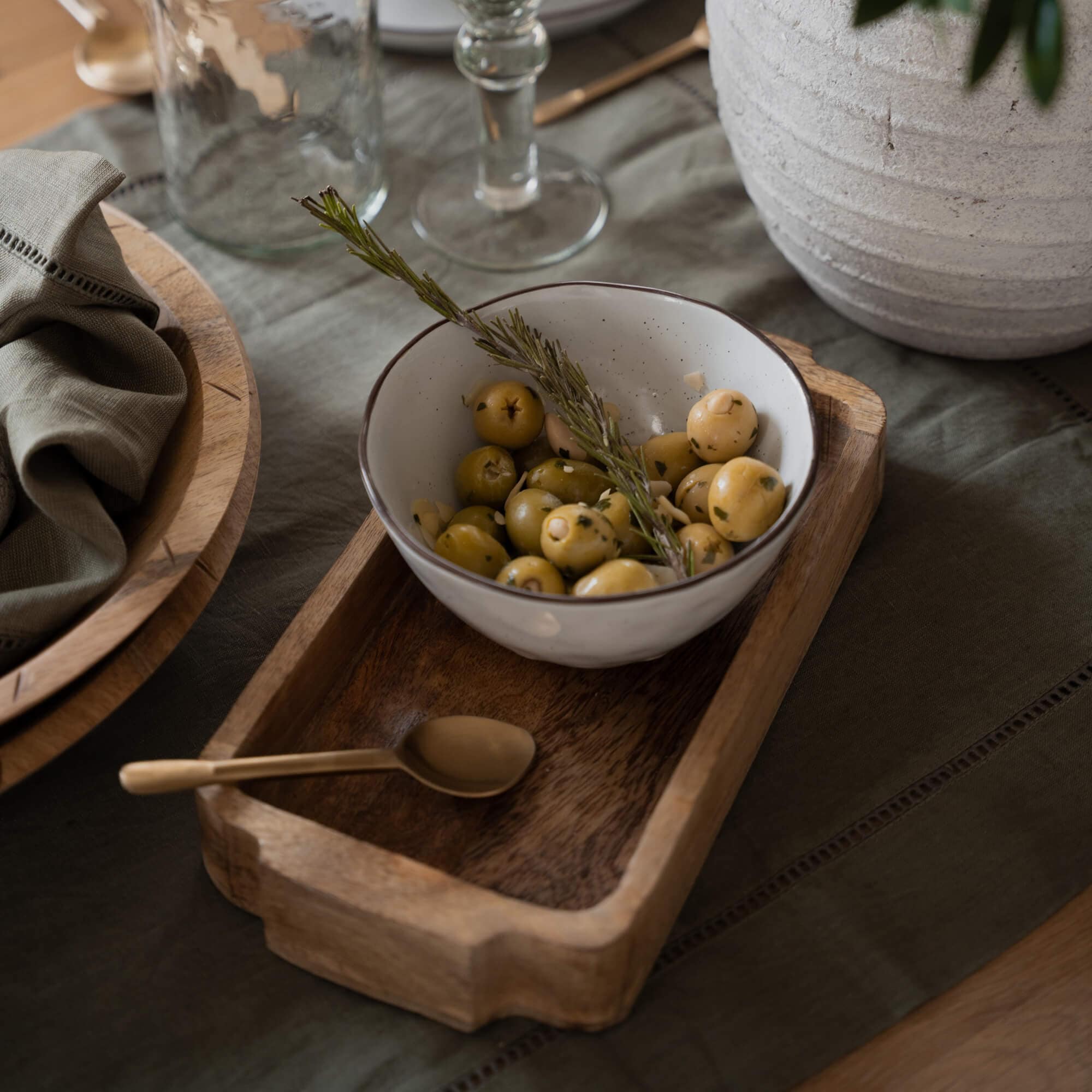 Bol en céramique blanche avec olives vertes et branche de romarin sur plateau en bois sur table
