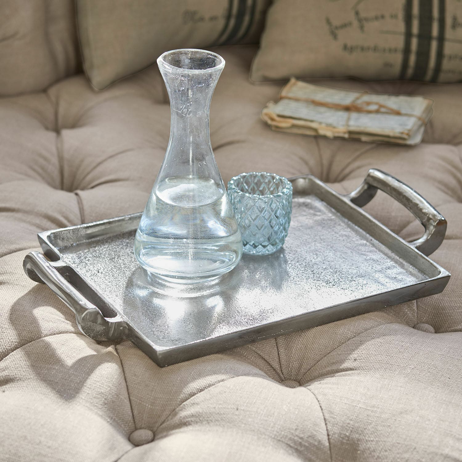 Plateau Connaire Plateau décoratif en métal argenté posé sur un tissu beige matelassé avec carafe et verre en verre