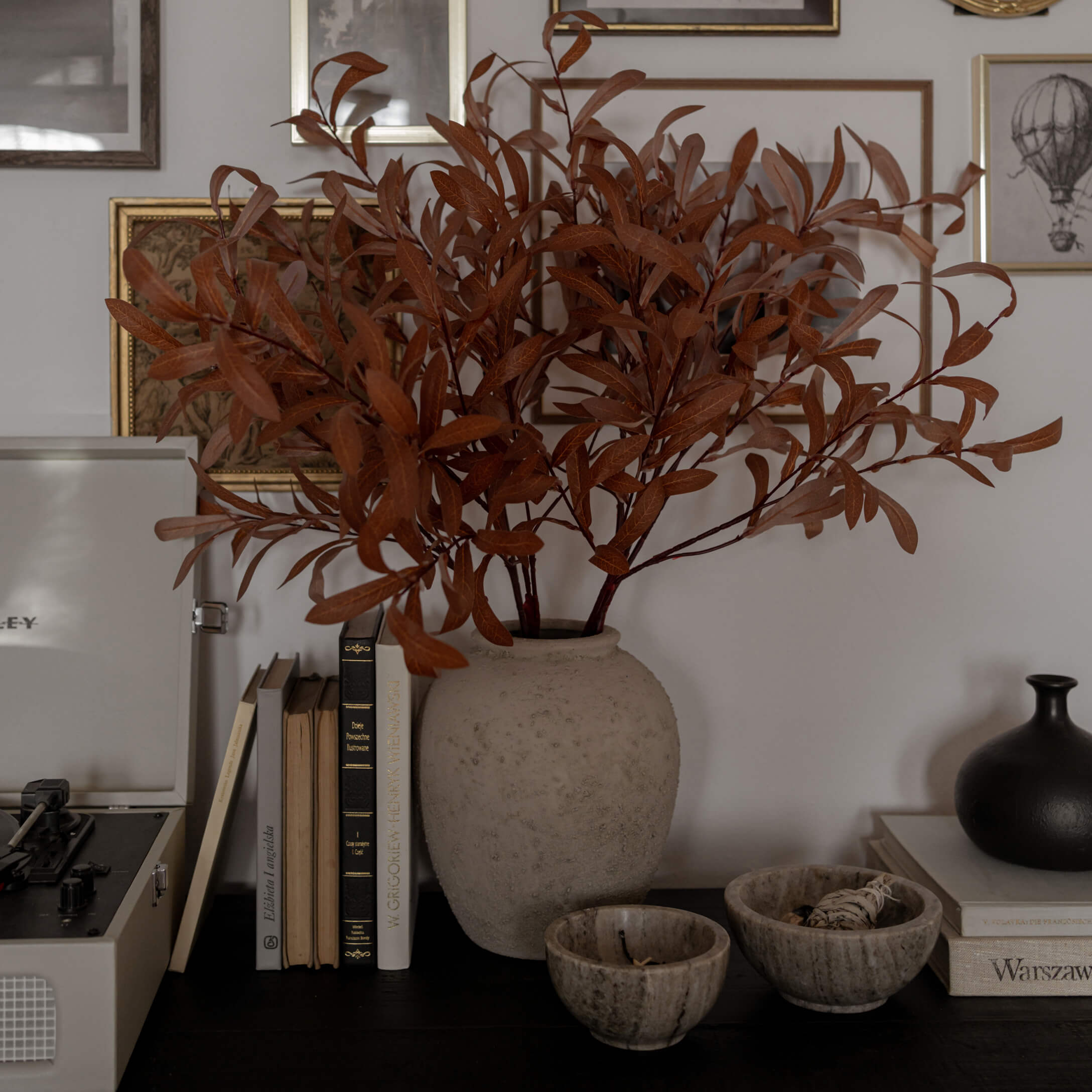 Vase en albâtre beige avec branches séchées brunes sur une table sombre avec livres et objets décoratifs