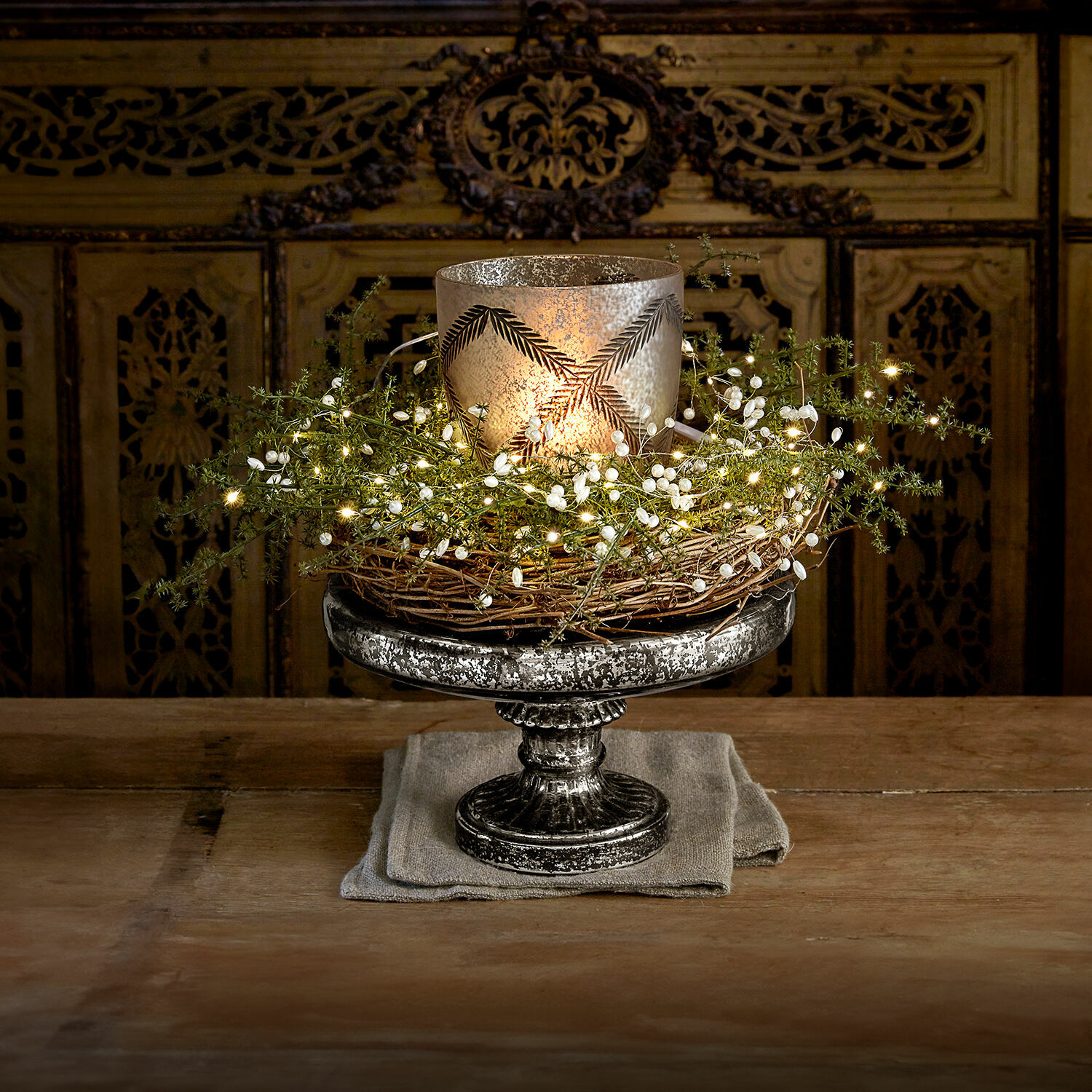 Présentoir à étage Incantesse Bougeoir décoratif en métal argenté avec couronne en osier et guirlande lumineuse LED sur table en bois