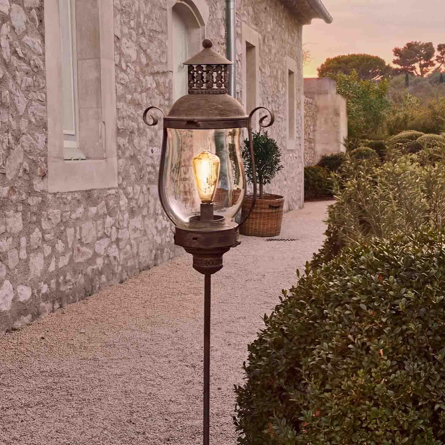 Lanterne de jardin en métal style rétro avec abat-jour transparent et ampoule LED devant une maison en pierre