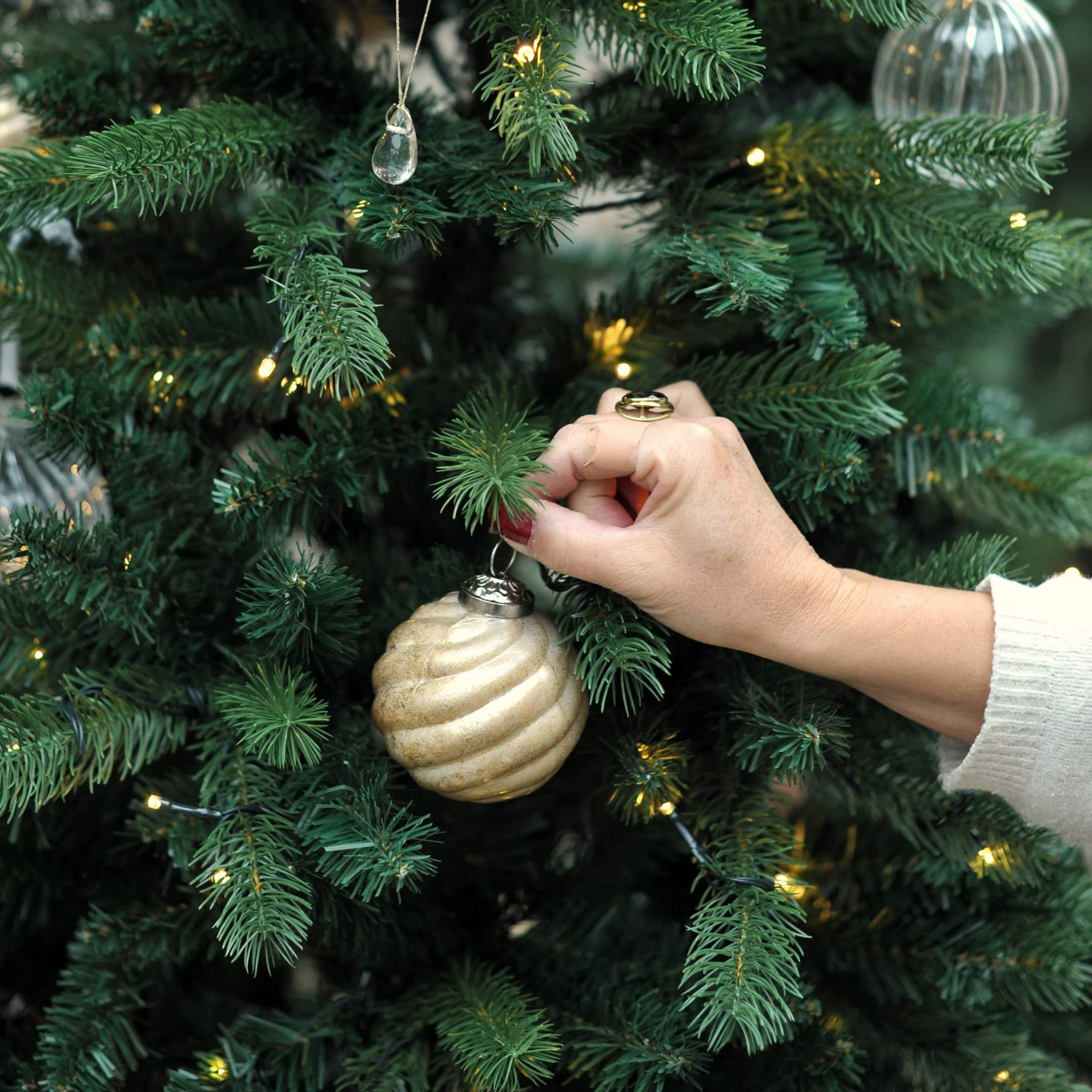 Main décorant un sapin vert avec une boule dorée spirale et des lumières LED dans une véranda