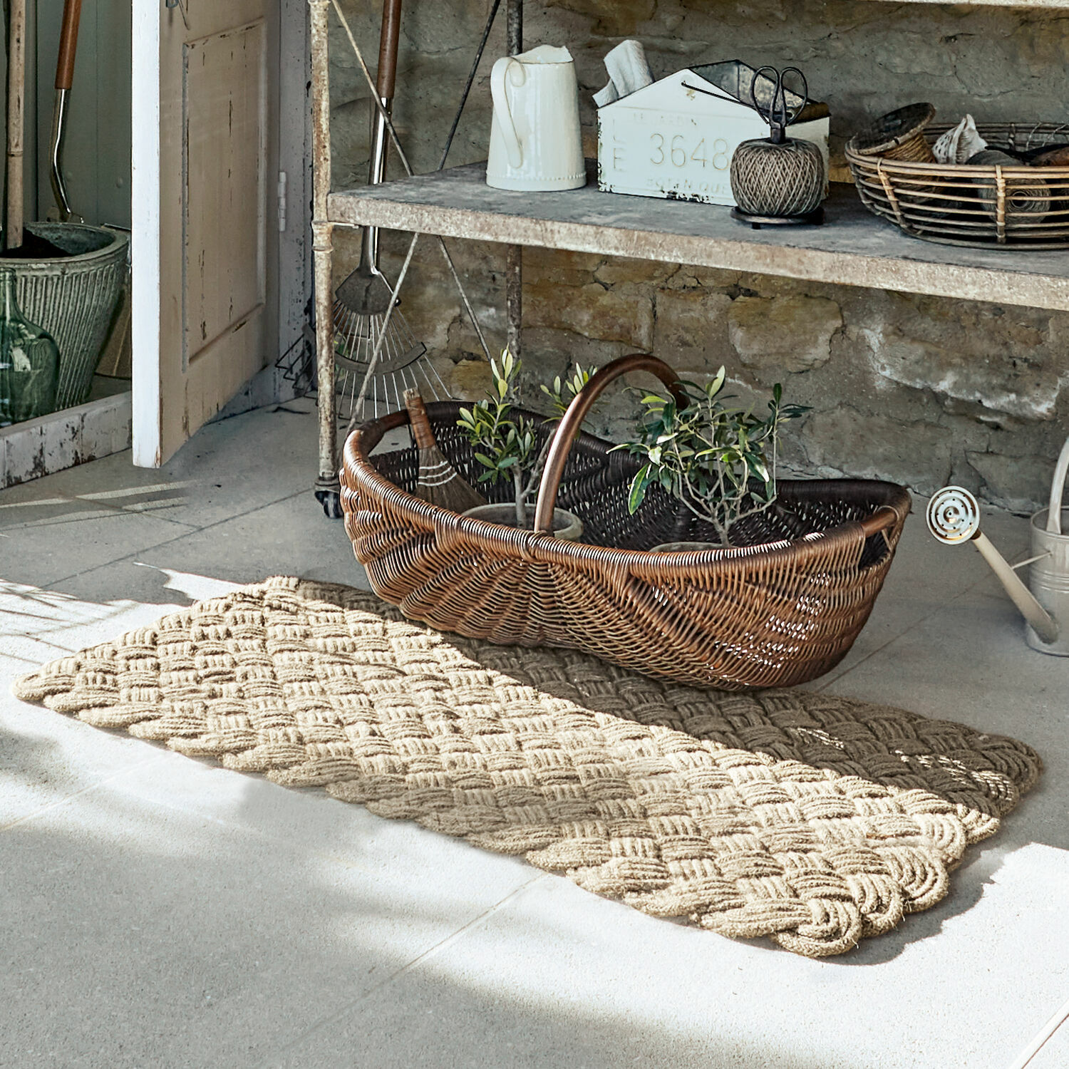 Tapis long Hildas Tapis beige tressé posé au sol dans un intérieur rustique avec panier en osier et outils de jardinage