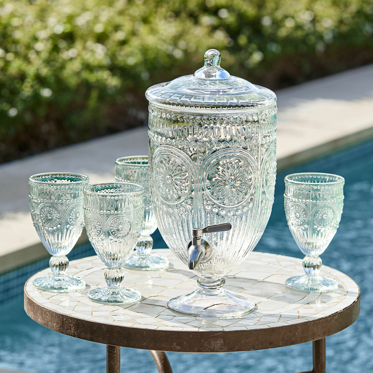 Distributeur de boisson Mercure Distributeur de boissons en verre avec couvercle et robinet, accompagné de quatre verres assortis sur une table près de la piscine