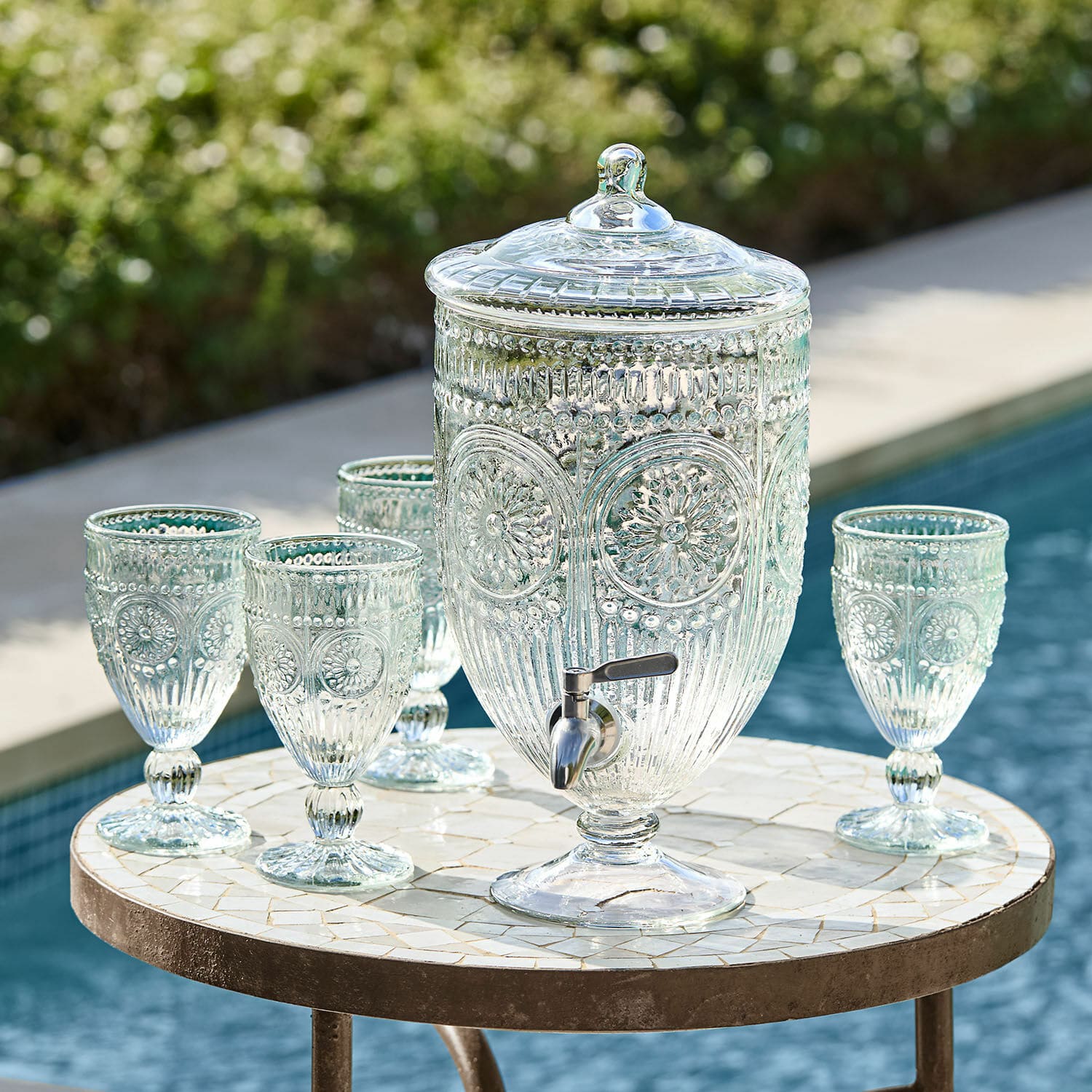 Distributeur de boisson Mercure Distributeur de boissons en verre avec couvercle et robinet, accompagné de quatre verres assortis sur une table près de la piscine