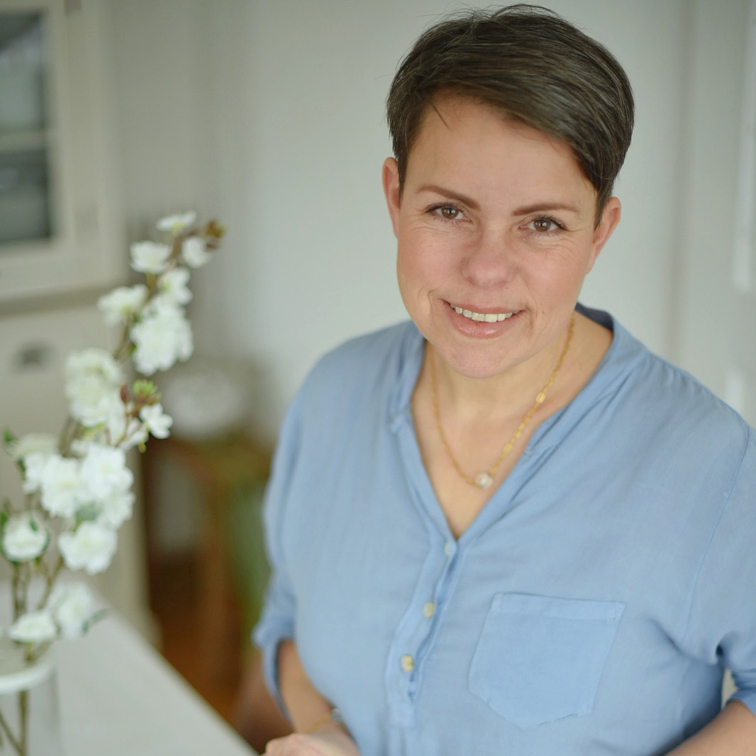 Femme souriante en chemise bleue près de fleurs blanches dans un intérieur lumineux