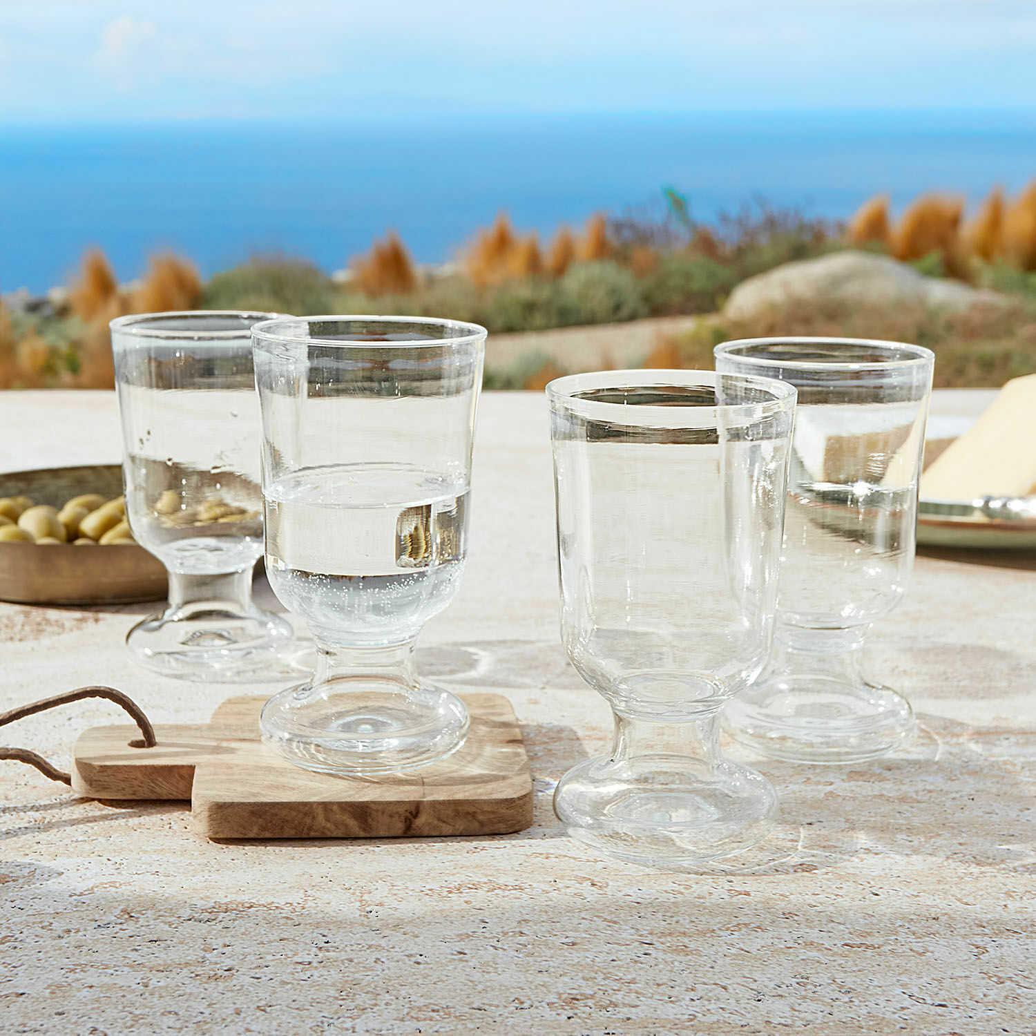 Lot de 4 verres Yvoncourt Ensemble de quatre verres transparents sur pied posés sur une table avec vue sur la mer