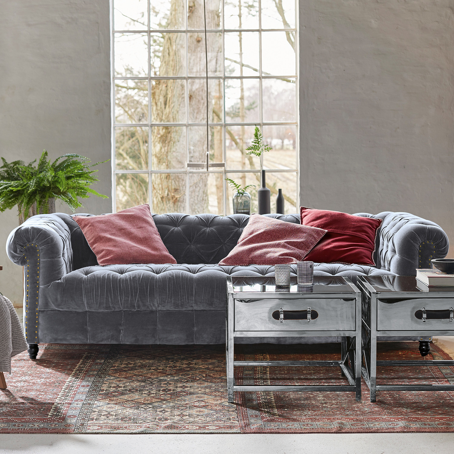 Canapé Chesterfield en velours gris avec coussins rouges et tables basses métalliques sur tapis décoratif