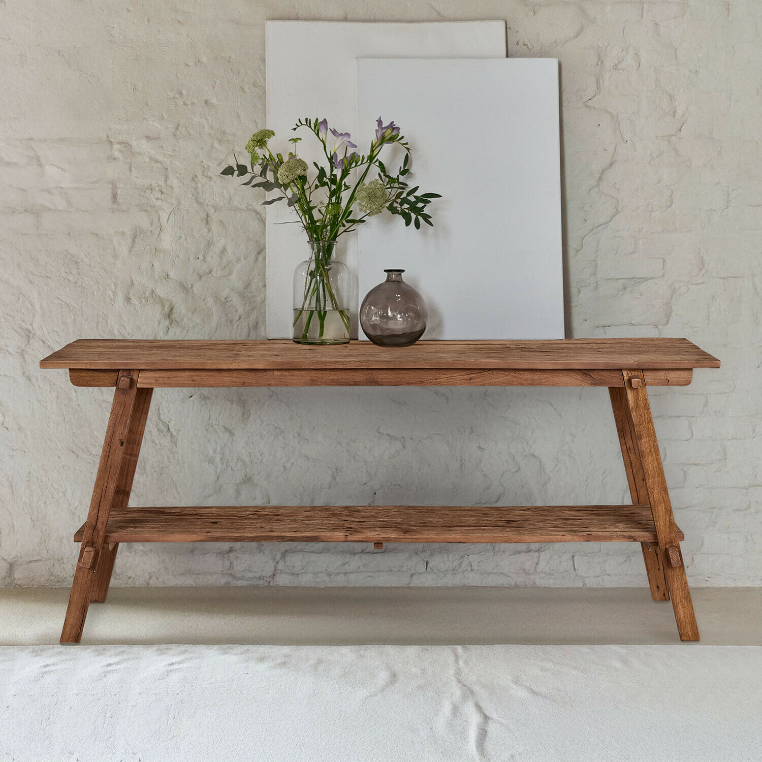 Console Brévonne Table en bois naturel avec étagère inférieure, décorée de vases et fleurs devant un mur blanc