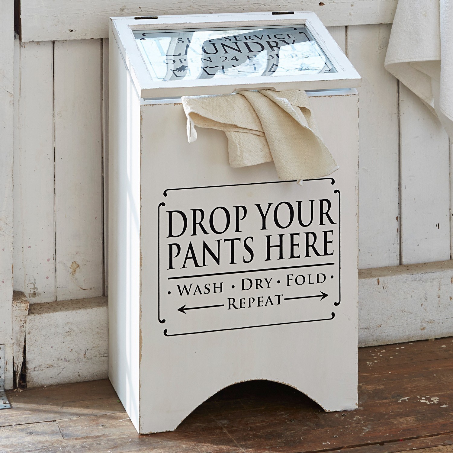 Coffre à linge Drop it Boîte à linge en bois blanc avec inscription et serviette crème posée sur un sol en bois