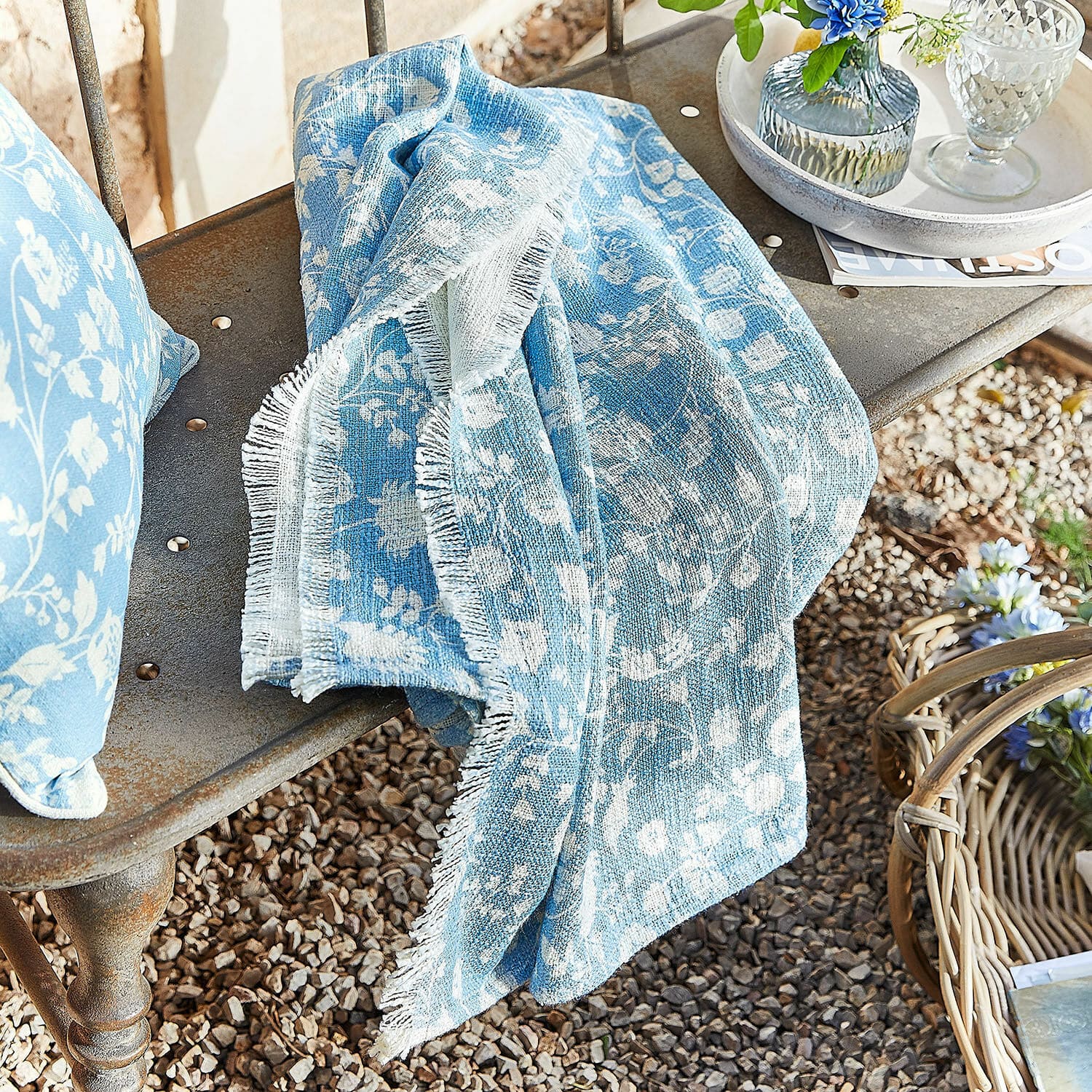Plaid Corvelique Plaid en coton bleu à motif floral blanc posé sur un banc en métal dans un jardin