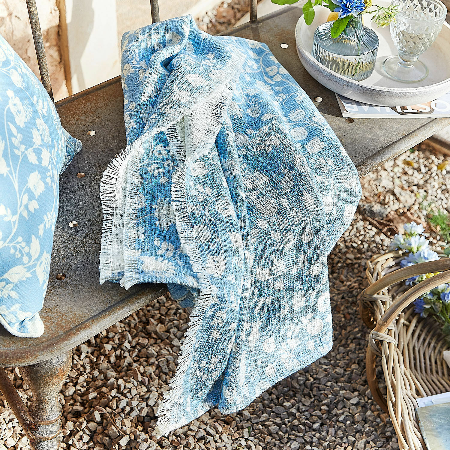 Plaid Corvelique Plaid en coton bleu à motif floral blanc posé sur un banc en métal dans un jardin