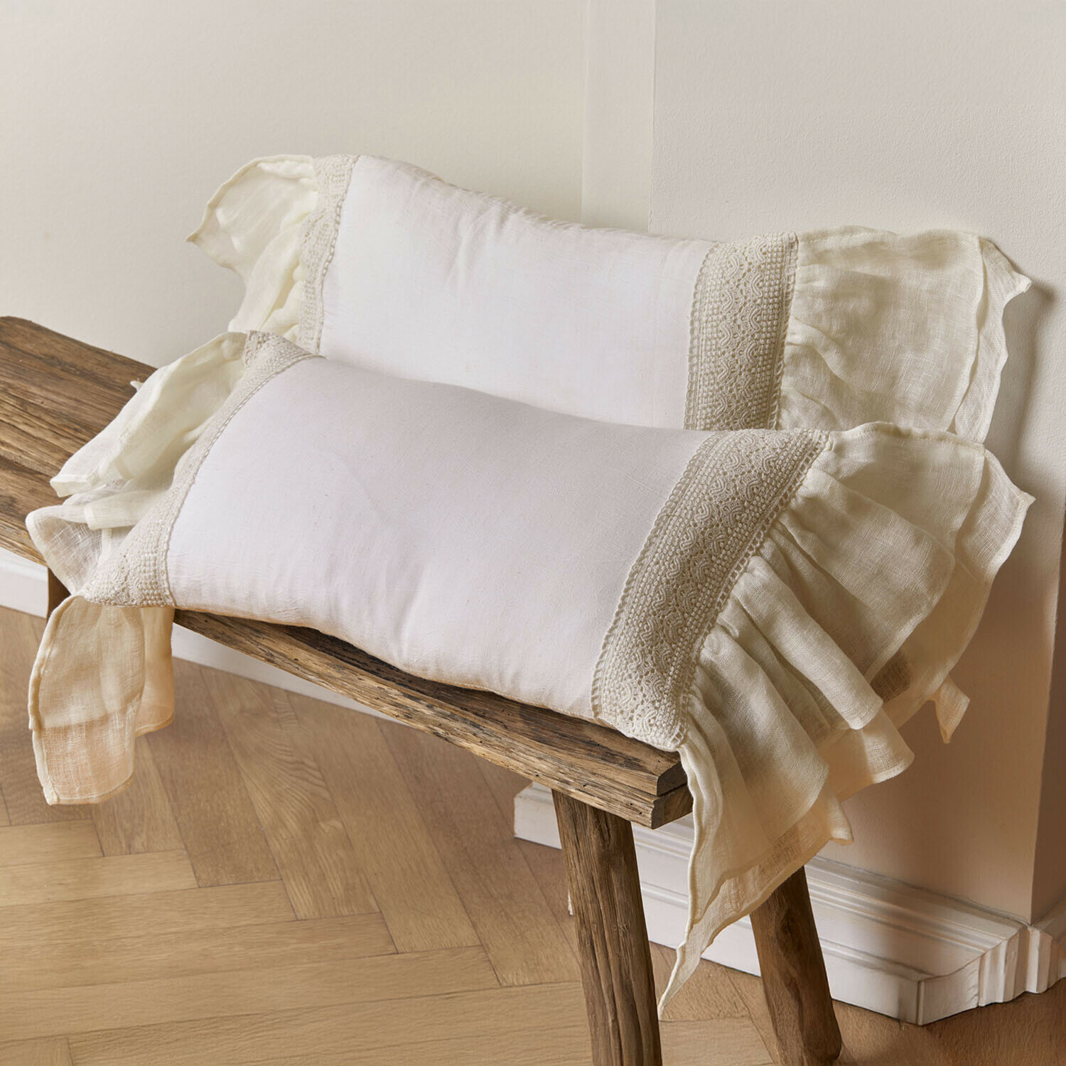 Lot de 2 housses de coussins Jolévianne Deux coussins en lin crème avec dentelle et volants posés sur un banc en bois