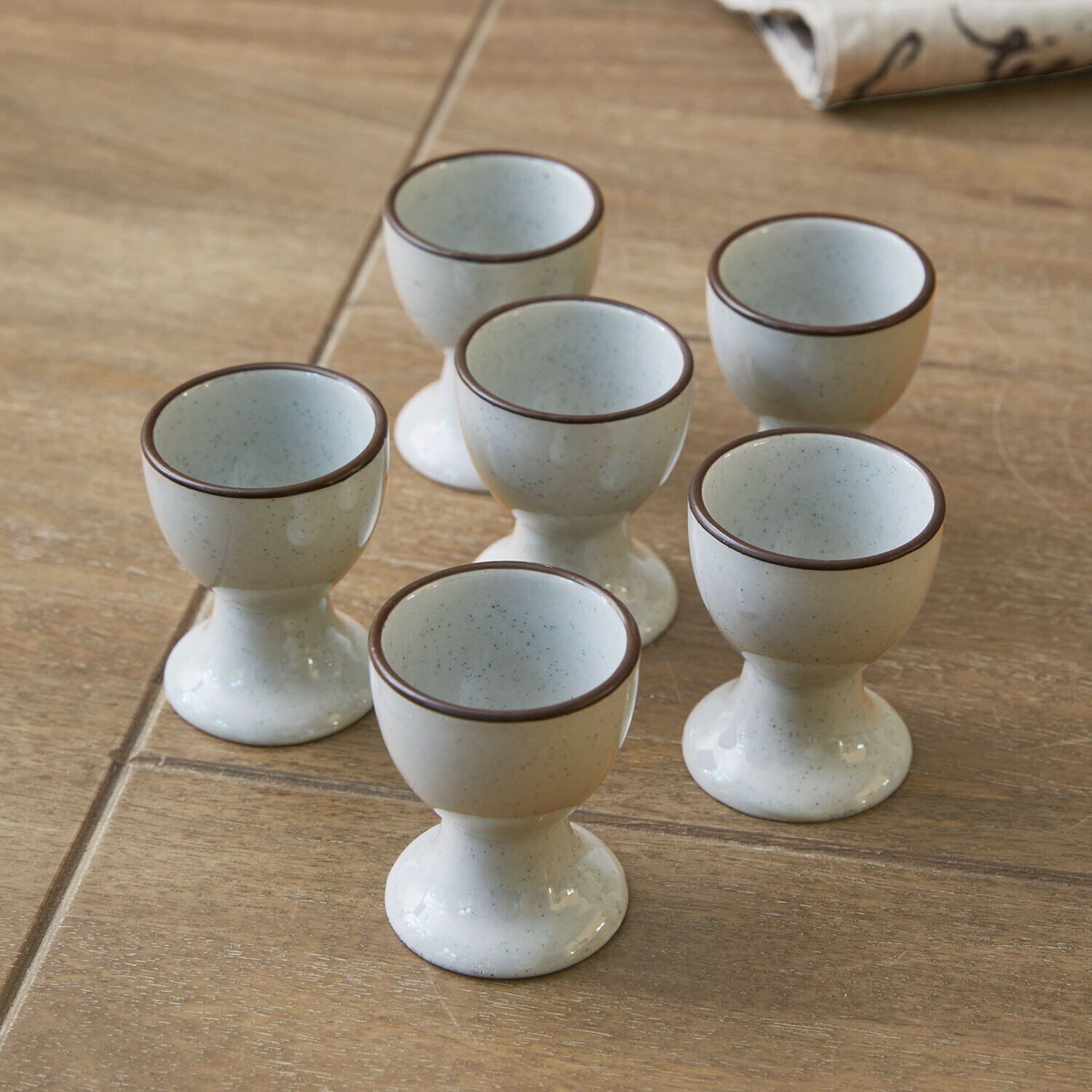 Lot de 6 coquetiers Biarré Ensemble de six coquetiers en céramique crème avec bordure marron posés sur une table en bois