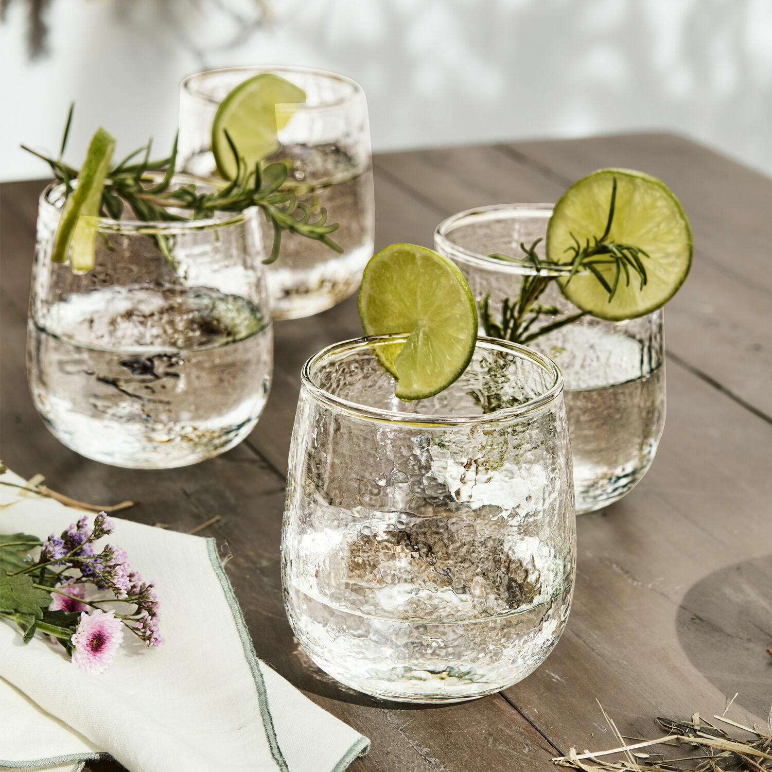 Lot de 4 verres Valters Ensemble de quatre verres en verre martelé avec tranches de citron vert et brins de romarin sur table en bois