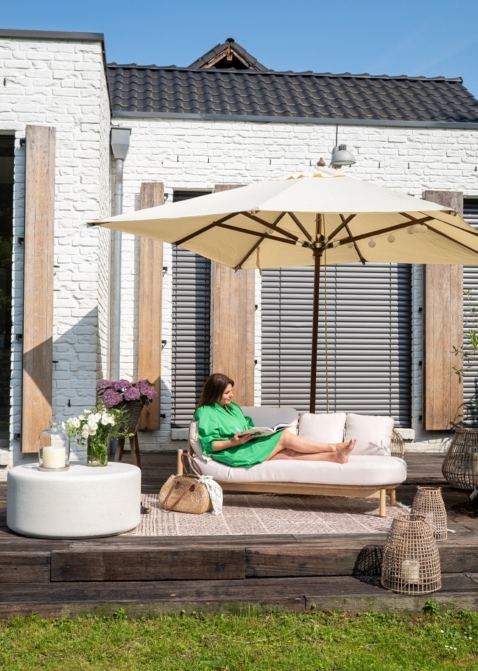 Femme en robe verte se détend sur un canapé clair sous un parasol crème sur une terrasse en bois