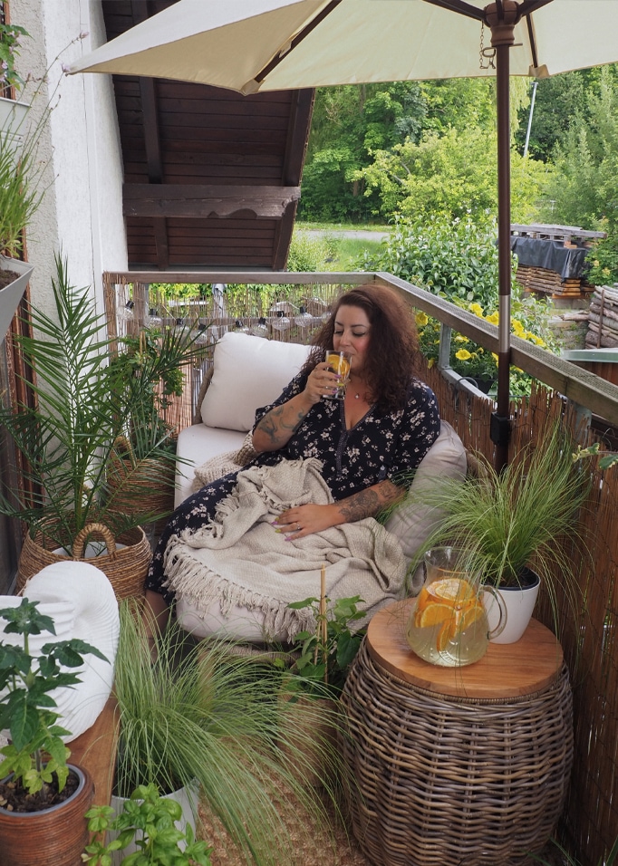 Femme se relaxant sur un balcon avec table en osier et couverture beige, entourée de plantes vertes.