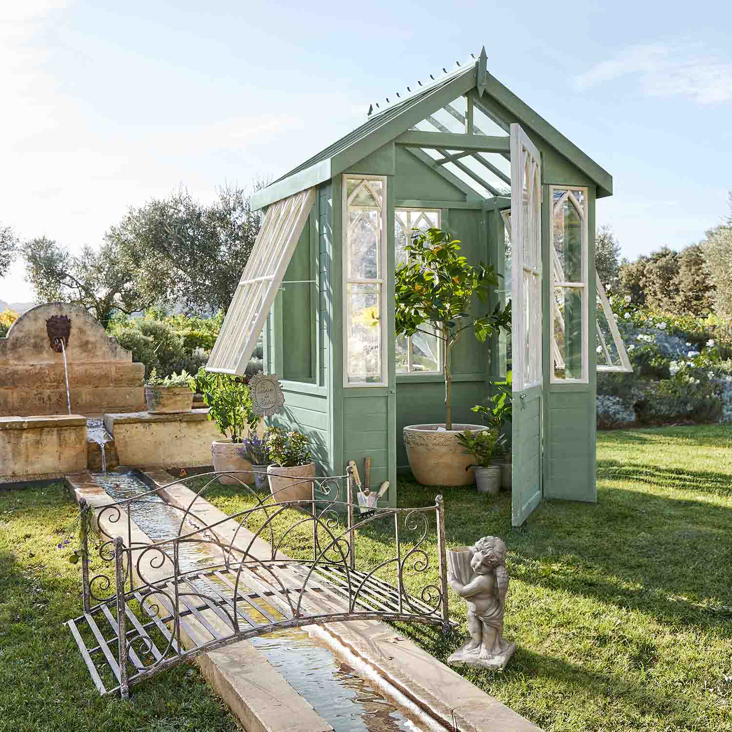 Serre en bois verte avec fenêtres blanches dans un jardin avec plantes et fontaine