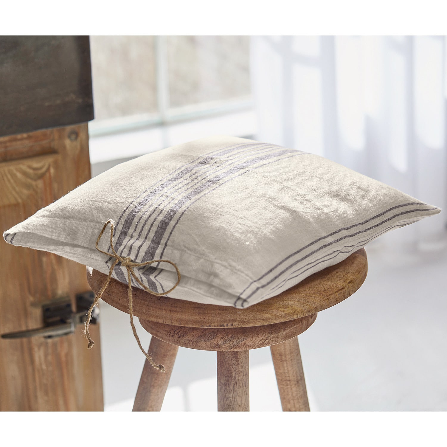 Coussin Amato Coussin décoratif en lin crème avec rayures grises posé sur un tabouret en bois