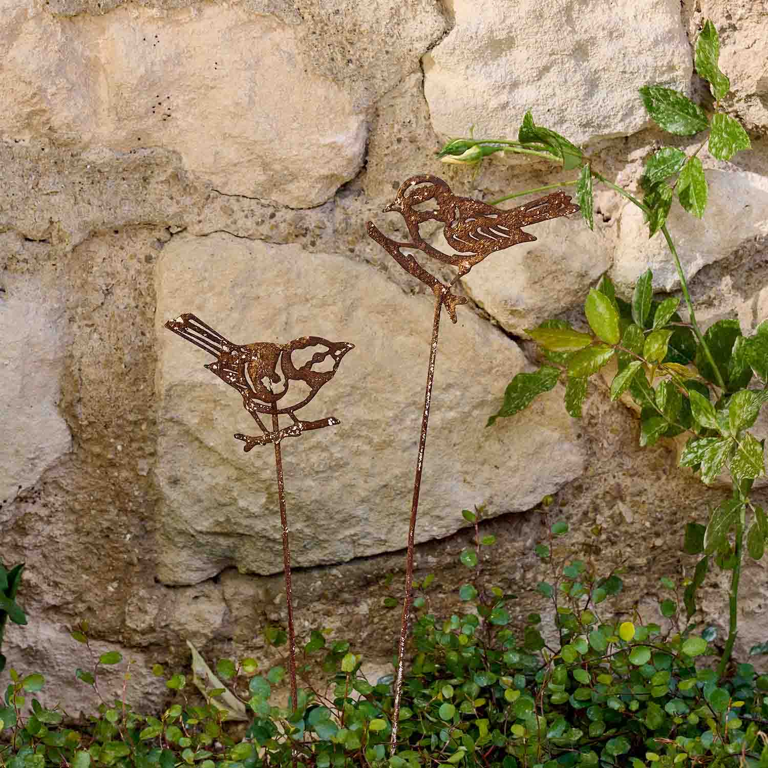 Oiseaux décoratifs en métal rouillé sur tiges dans un jardin devant un mur en pierre