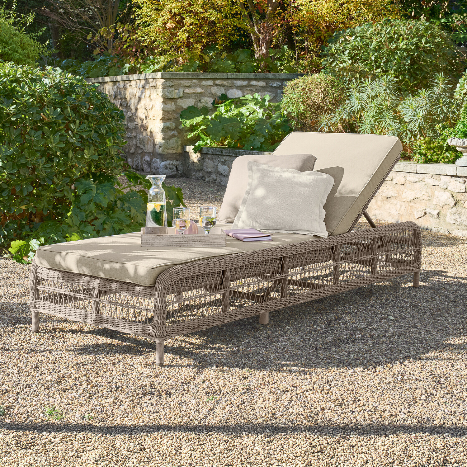 Chaise longue Hereford Chaise longue de jardin beige avec coussin et structure en osier sur sol en gravier dans un jardin verdoyant