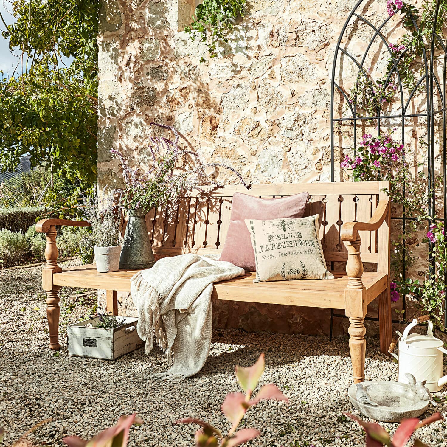 Banc Loudoun Banc de jardin en bois clair avec coussins et plaid sur patio en gravier entouré de plantes